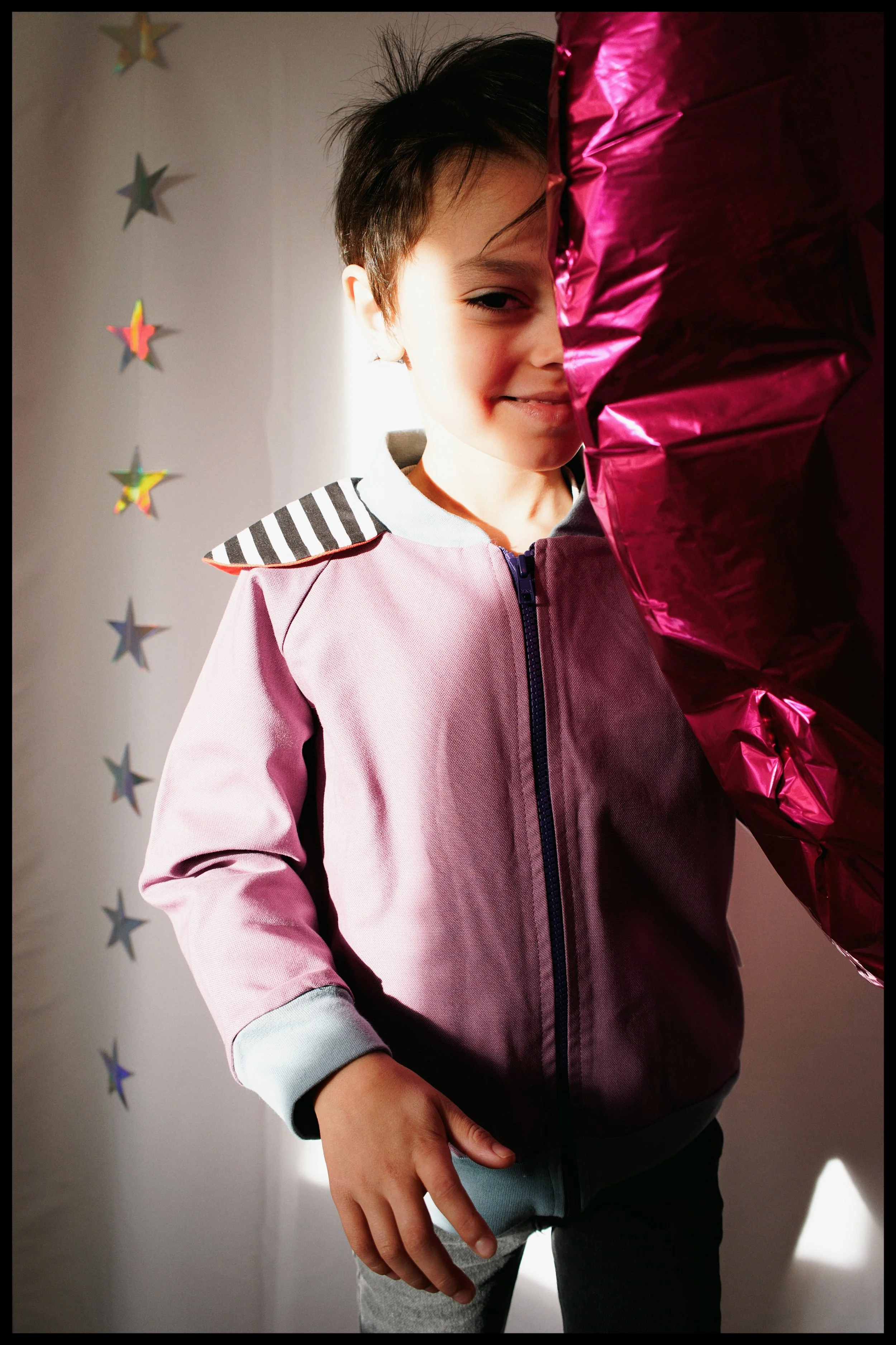 A young boy with dark hair smiling and holding a shiny pink balloon near his face. He wears a light pink jacket and dark pants, with star-shaped decorations on the white wall behind him.