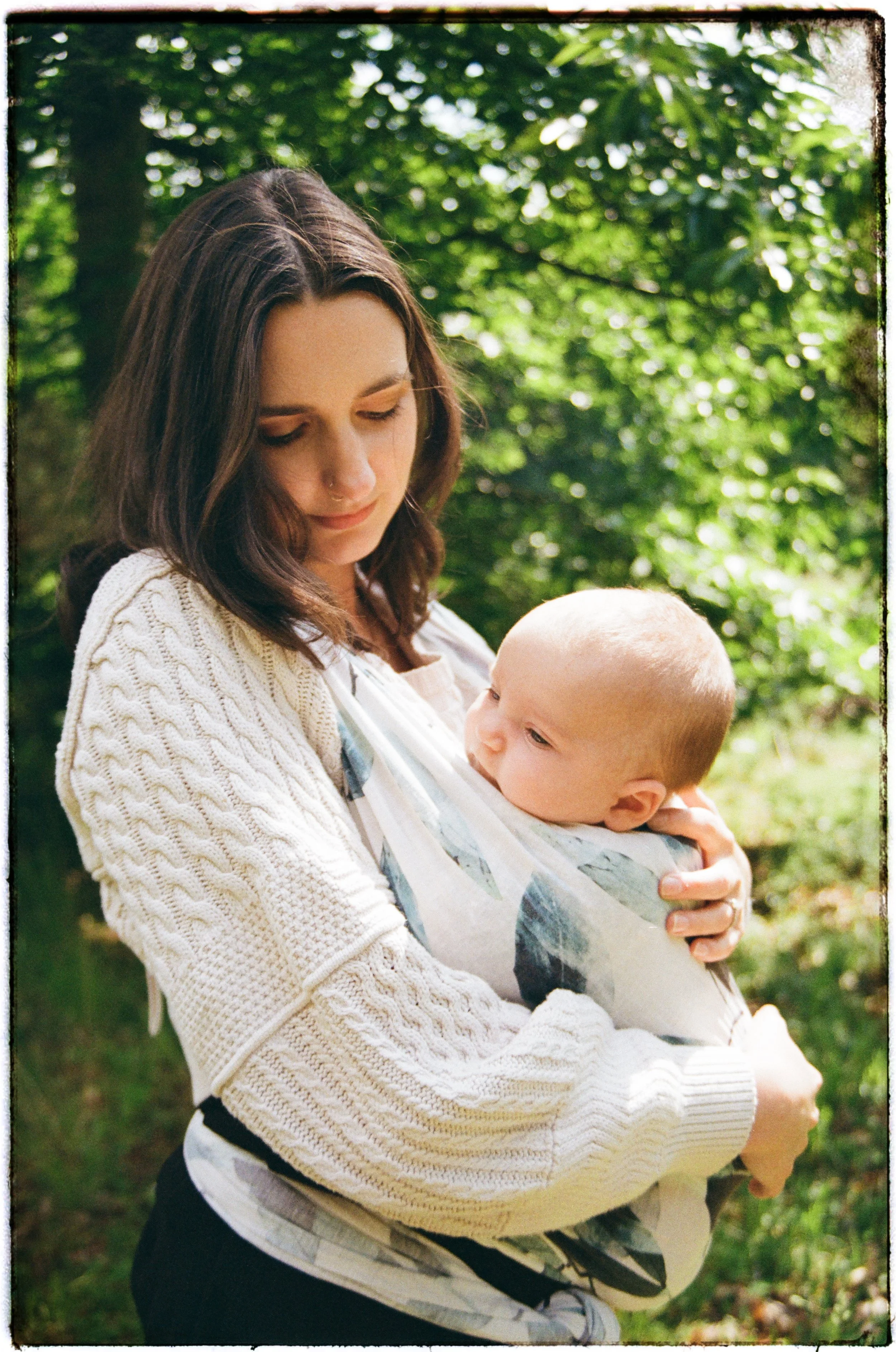 A woman holding a baby outdoors in a green, leafy environment, with both looking down. The woman has brown hair and is wearing a white knitted sweater. The baby is wrapped in a colorful blanket.