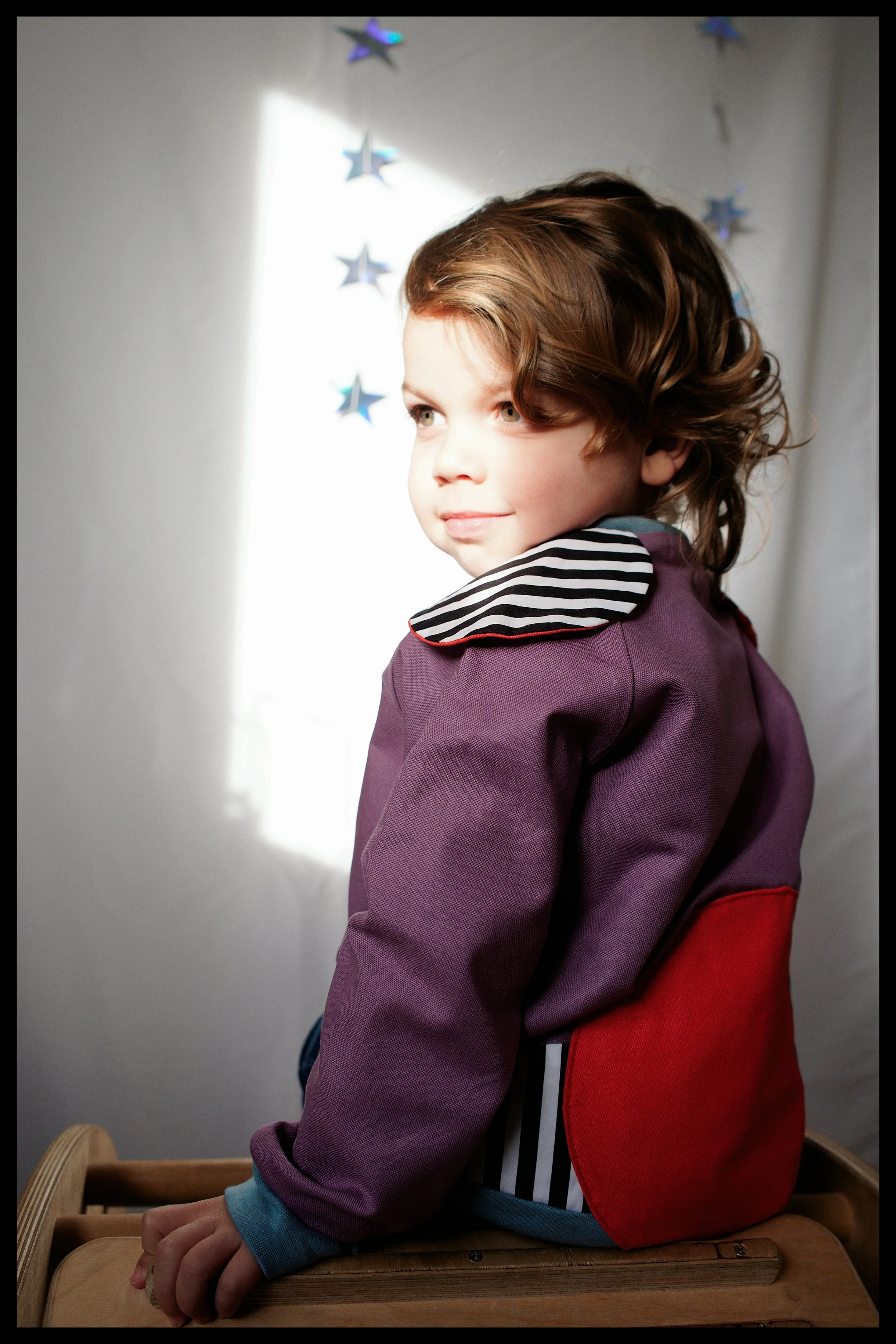 A young boy with curly brown hair and light-colored eyes sitting at a wooden table, wearing a purple jacket with a striped collar, and a red backpack. There are hanging star decorations in the background. Light is coming from a window, illuminating h