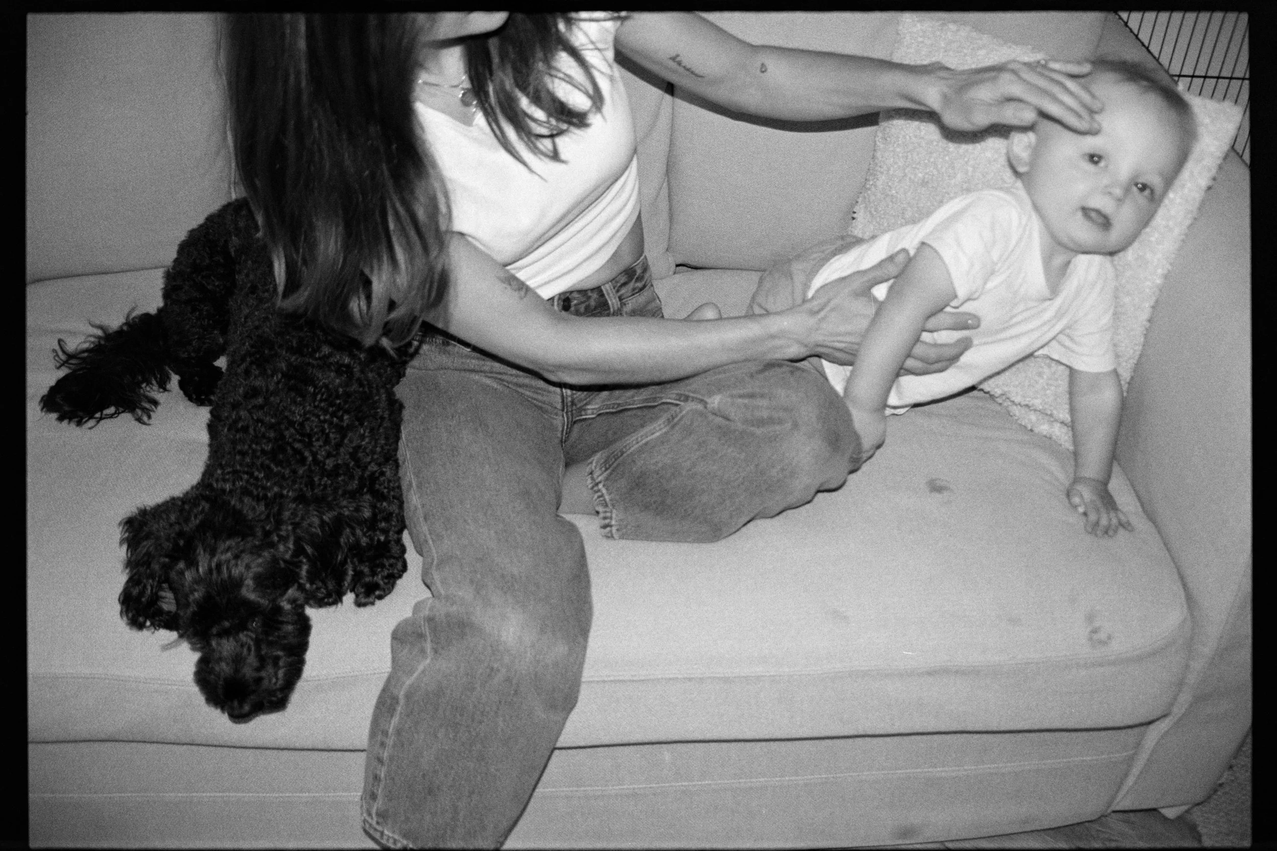 A woman with long hair, wearing jeans and a striped shirt, is sitting on a sofa next to a small child lying on their stomach on a textured blanket. A black dog is partially visible near the woman's lap, and the woman's hand is gently extended towards