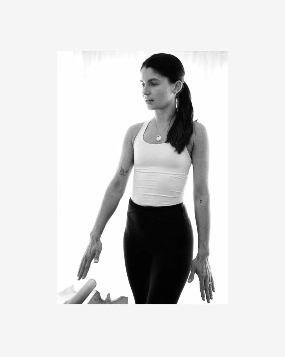 A young woman with long dark hair in a ponytail, wearing a white tank top and black pants, standing indoors with natural light behind her.