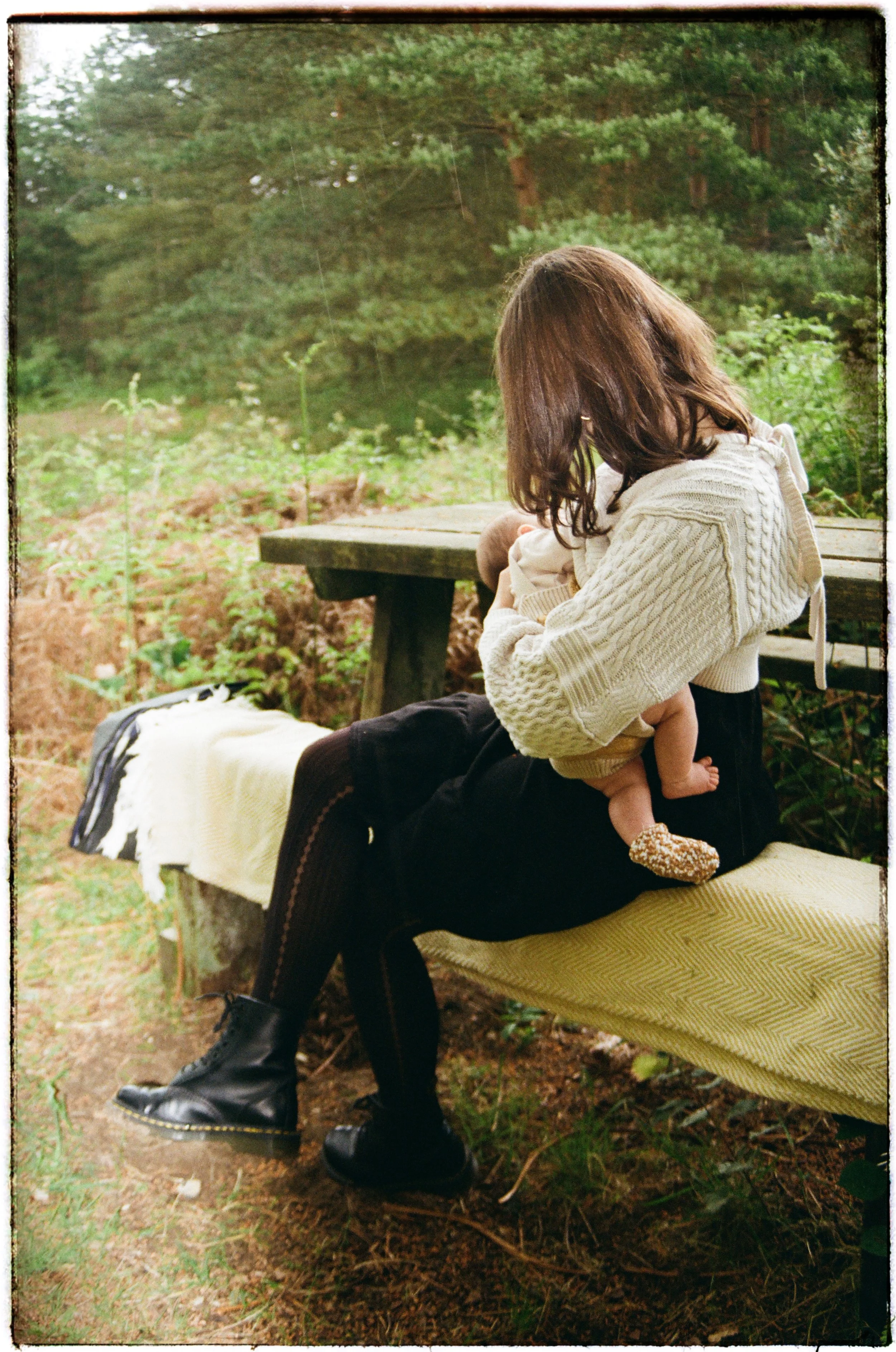 A woman sitting outdoors in a natural setting, holding a child, with a wooden table nearby and trees in the background.
