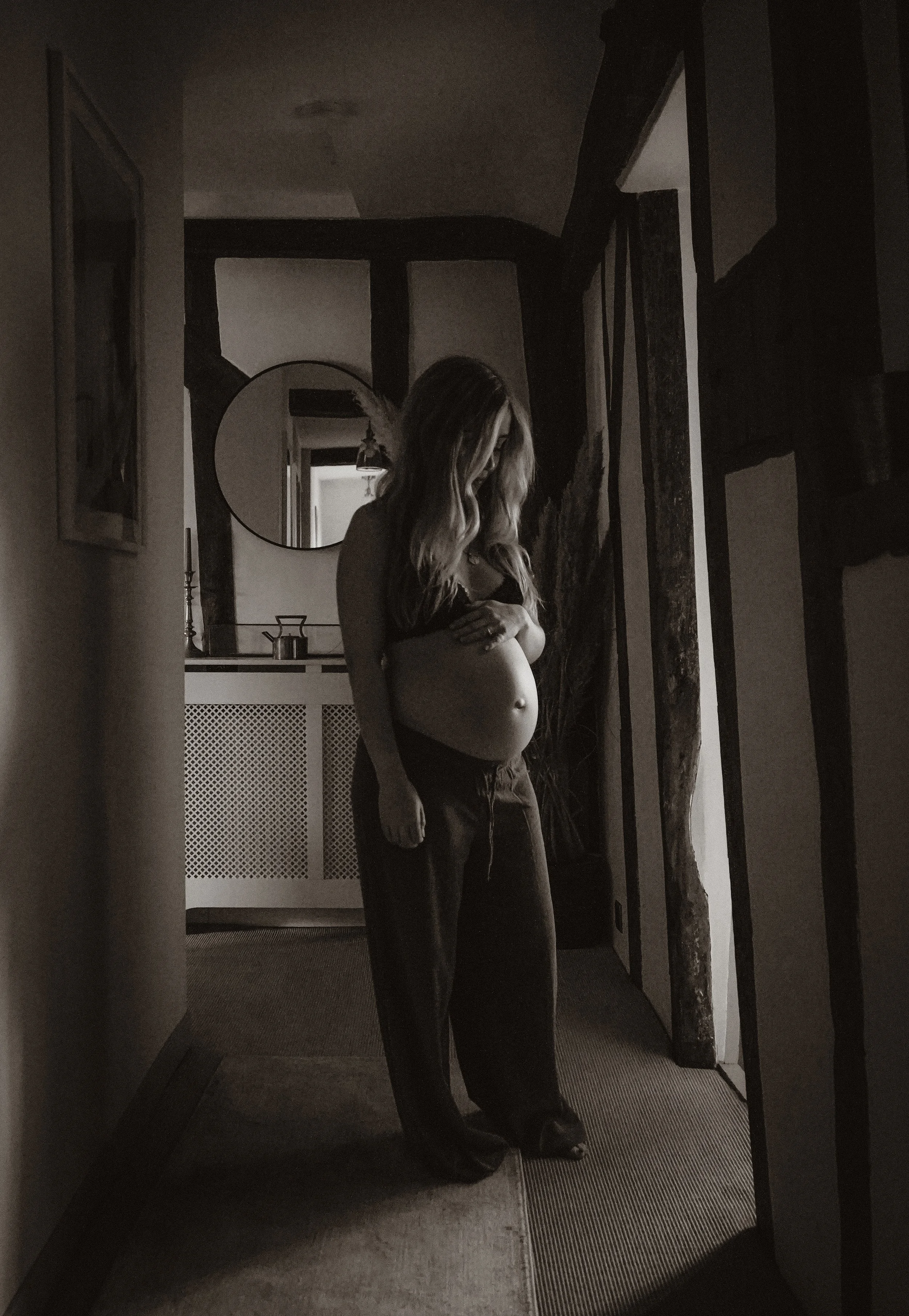 A woman standing in a hallway, holding her chest and looking down, with a mirror and decorative objects visible in the background, in black and white.
