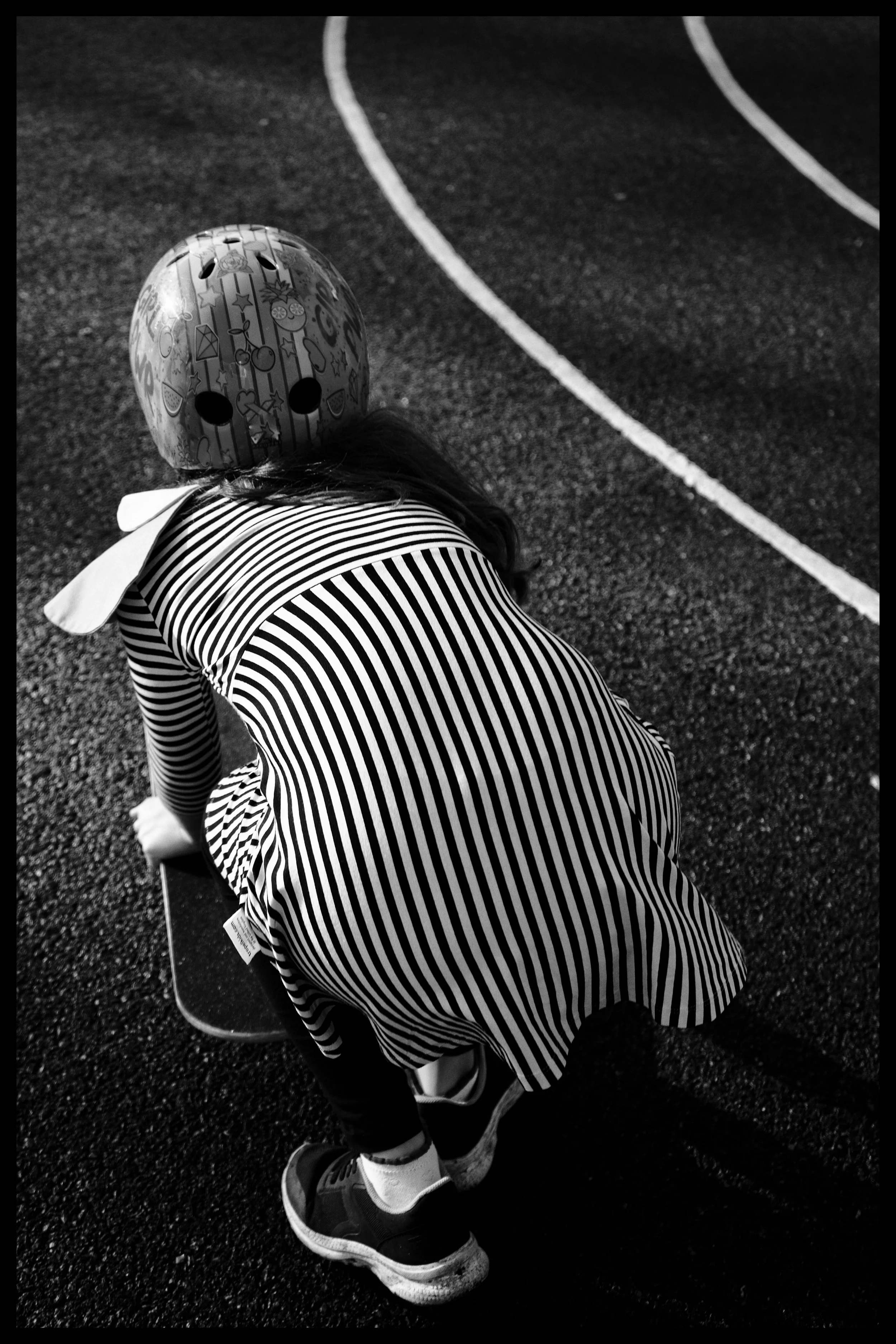 Child in striped dress and sneakers bending over on a running track, holding a skateboard, wearing a decorative helmet with emojis.