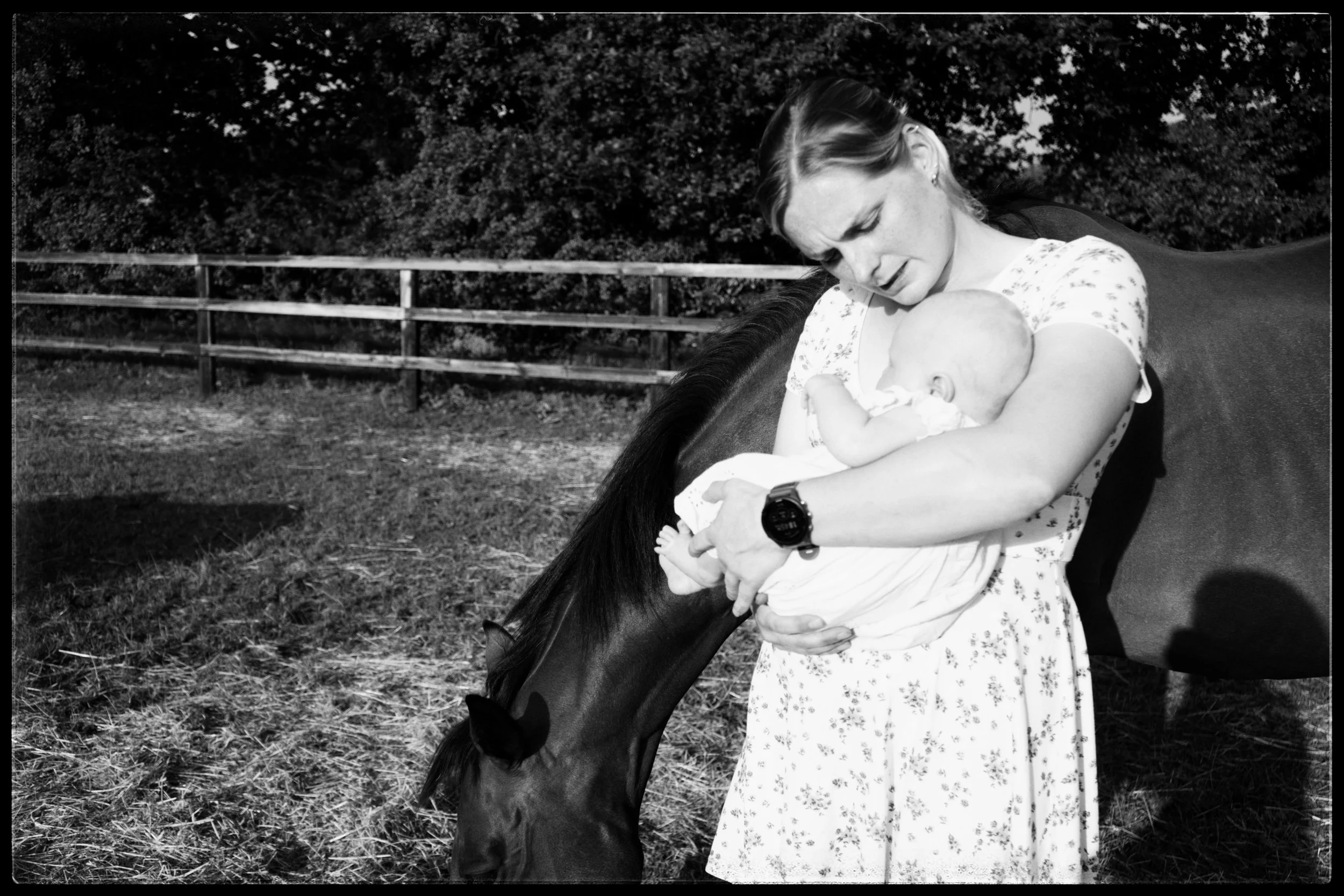 A woman is holding a young child while sitting on a horse outdoors. The background features a wooden fence and trees.