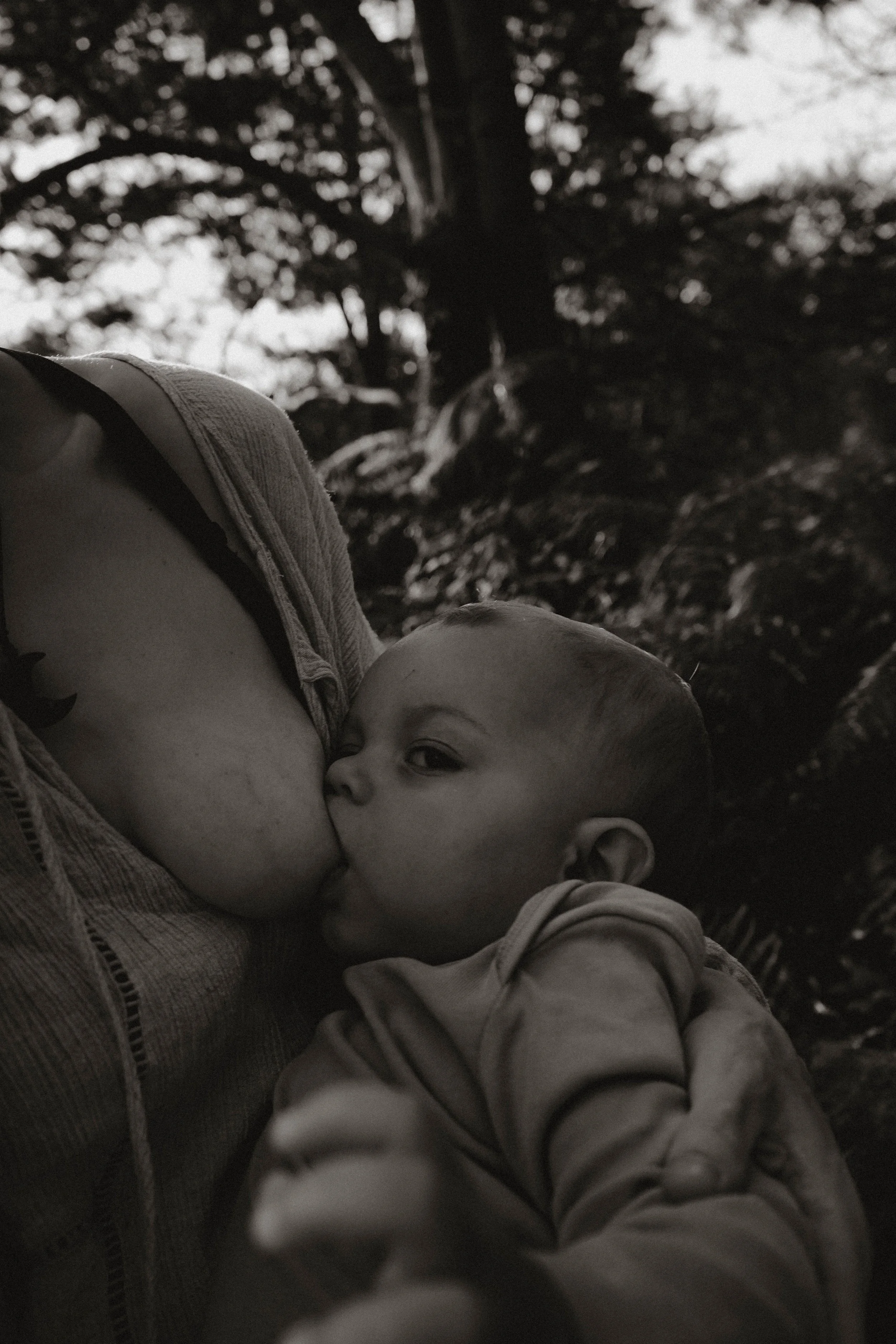 A black and white photo of a person holding a sleeping baby close, with trees in the background.