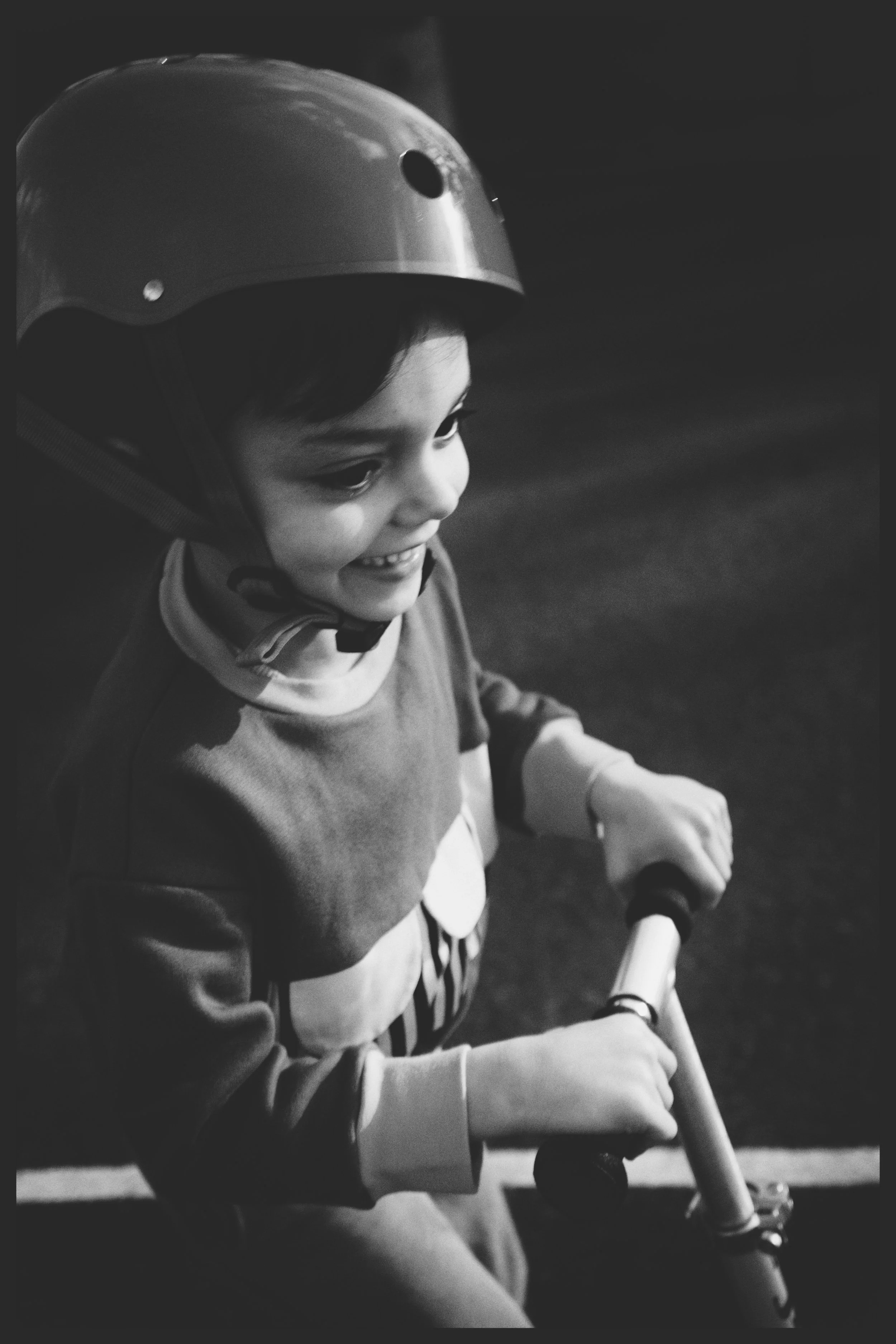 A young child wearing a helmet is riding a scooter indoors, smiling and looking to the side.