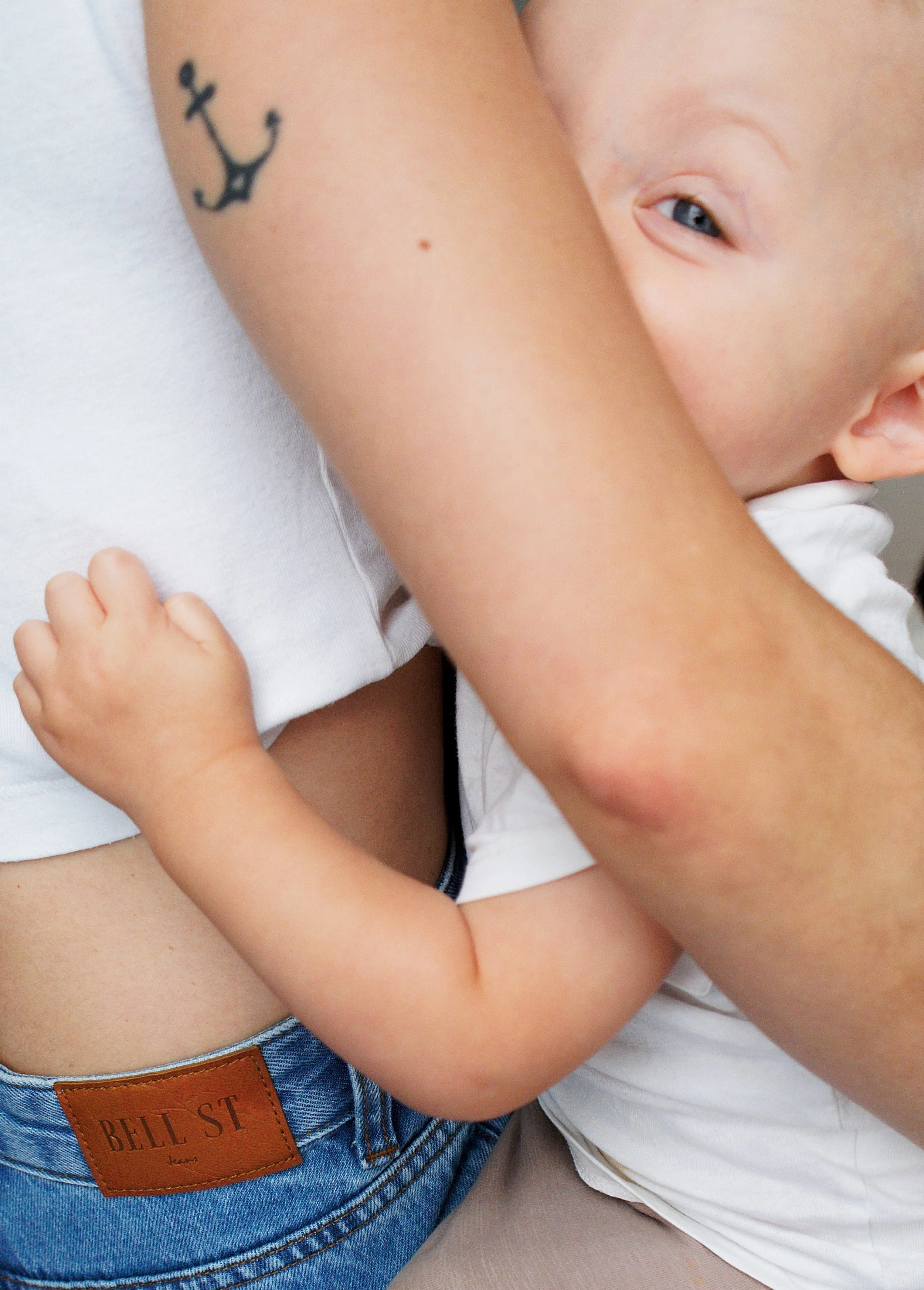 Close-up of a child's arms hugging an adult, close to their face showing a partial view of the child's eye and a tattoo of an anchor on the child's arm, with the adult wearing a white shirt.