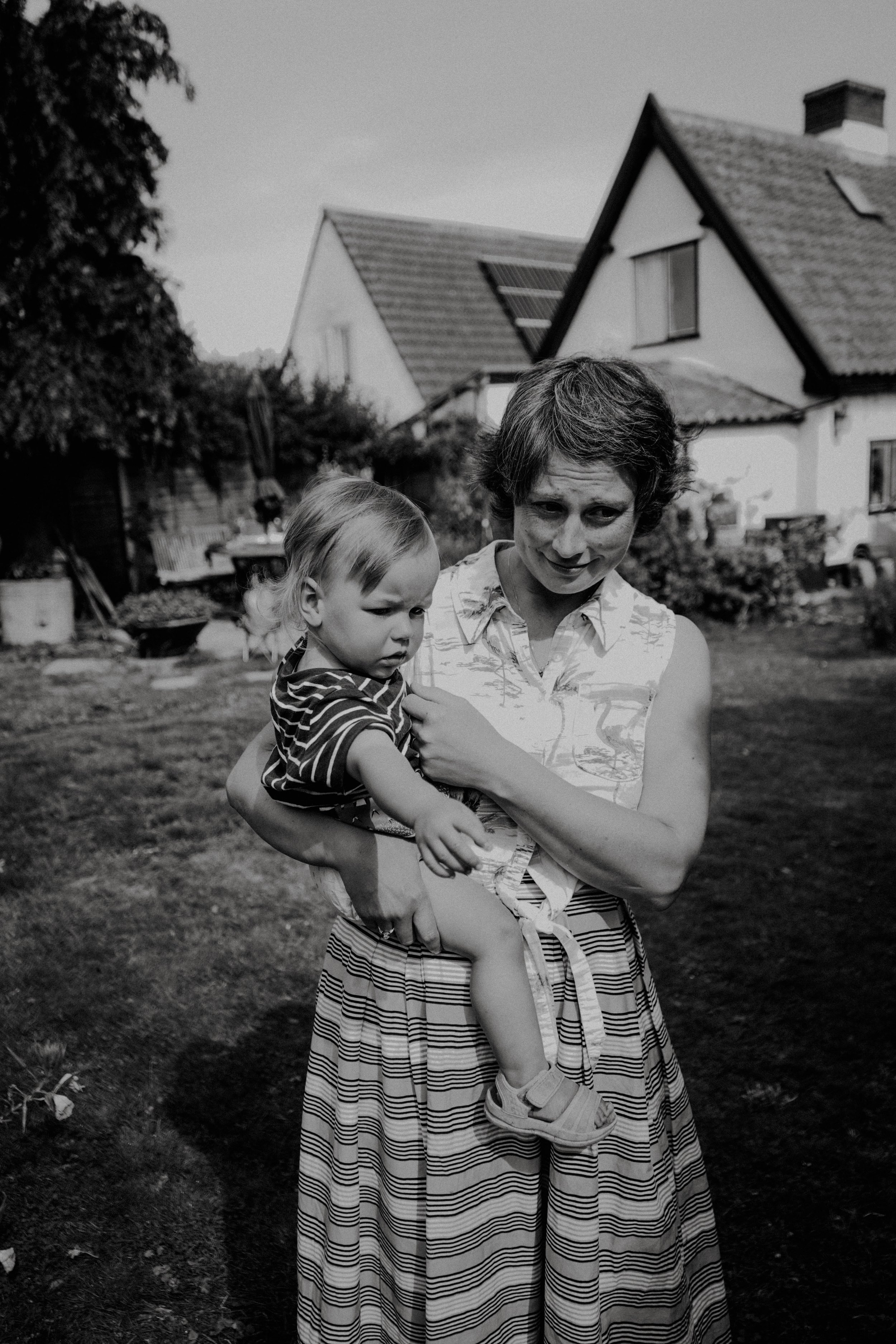 A woman holding a young child outdoors in a yard with a house in the background. The woman has short, curly hair and is wearing a striped skirt and sleeveless top. The child has light-colored hair and is wearing a striped shirt.