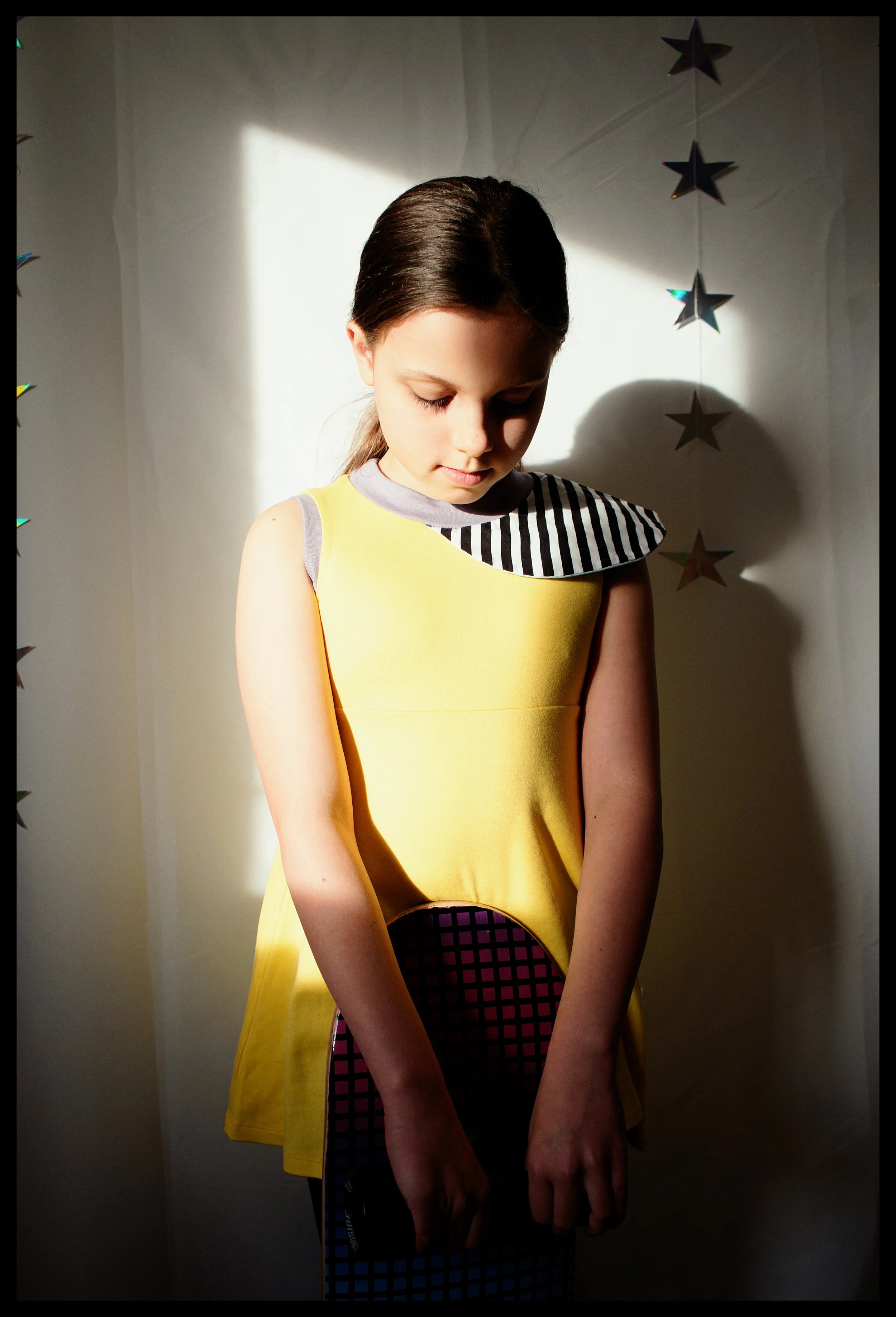A young girl with long brown hair in a ponytail wearing a sleeveless yellow dress with a black and white striped collar, standing in front of a plain light-colored wall with star-shaped decorations hanging on the right side. She is looking downward w