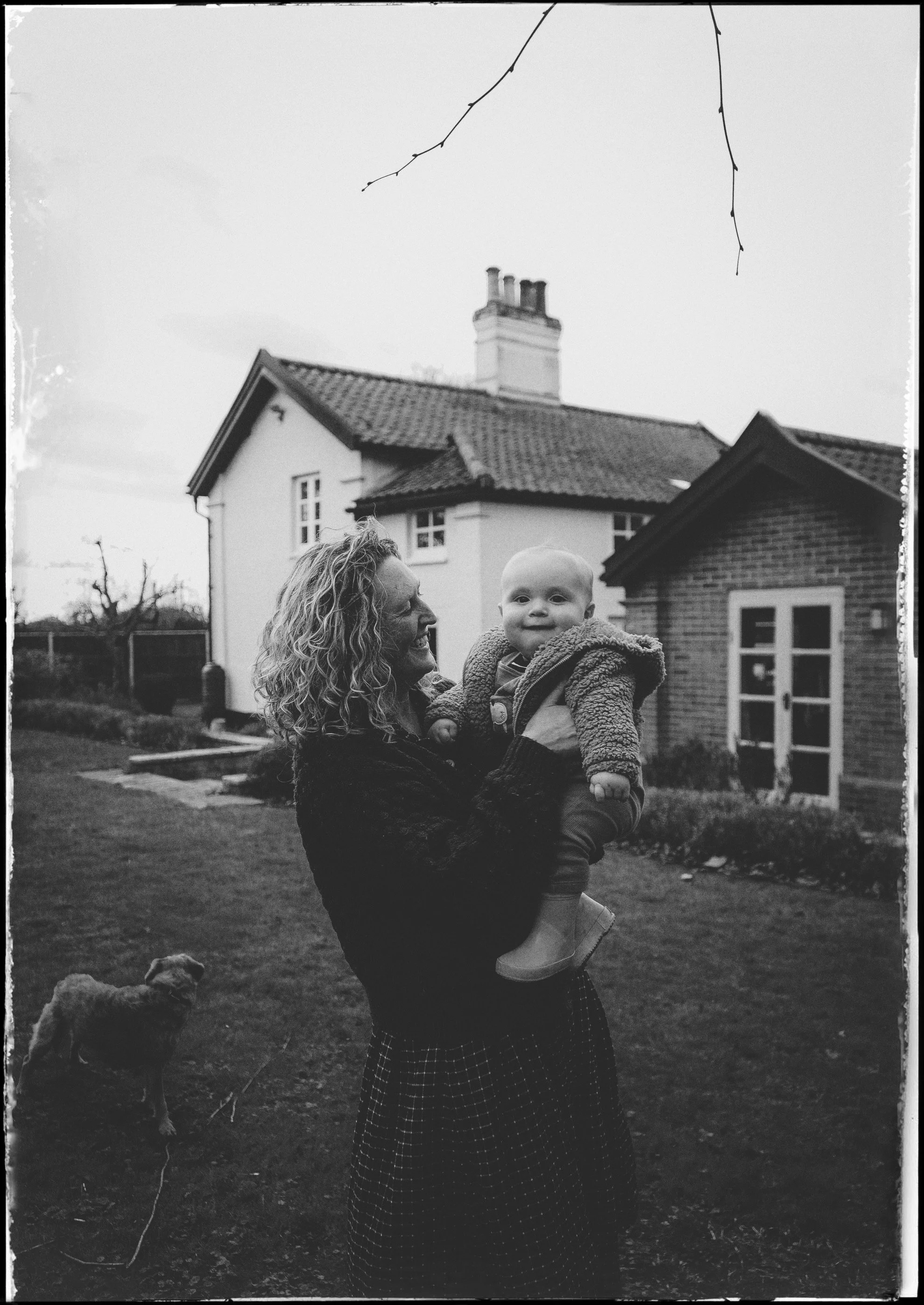 A woman with curly hair holding a smiling baby outdoors in a yard with houses in the background. There is a dog in the foreground and bare tree branches overhead.