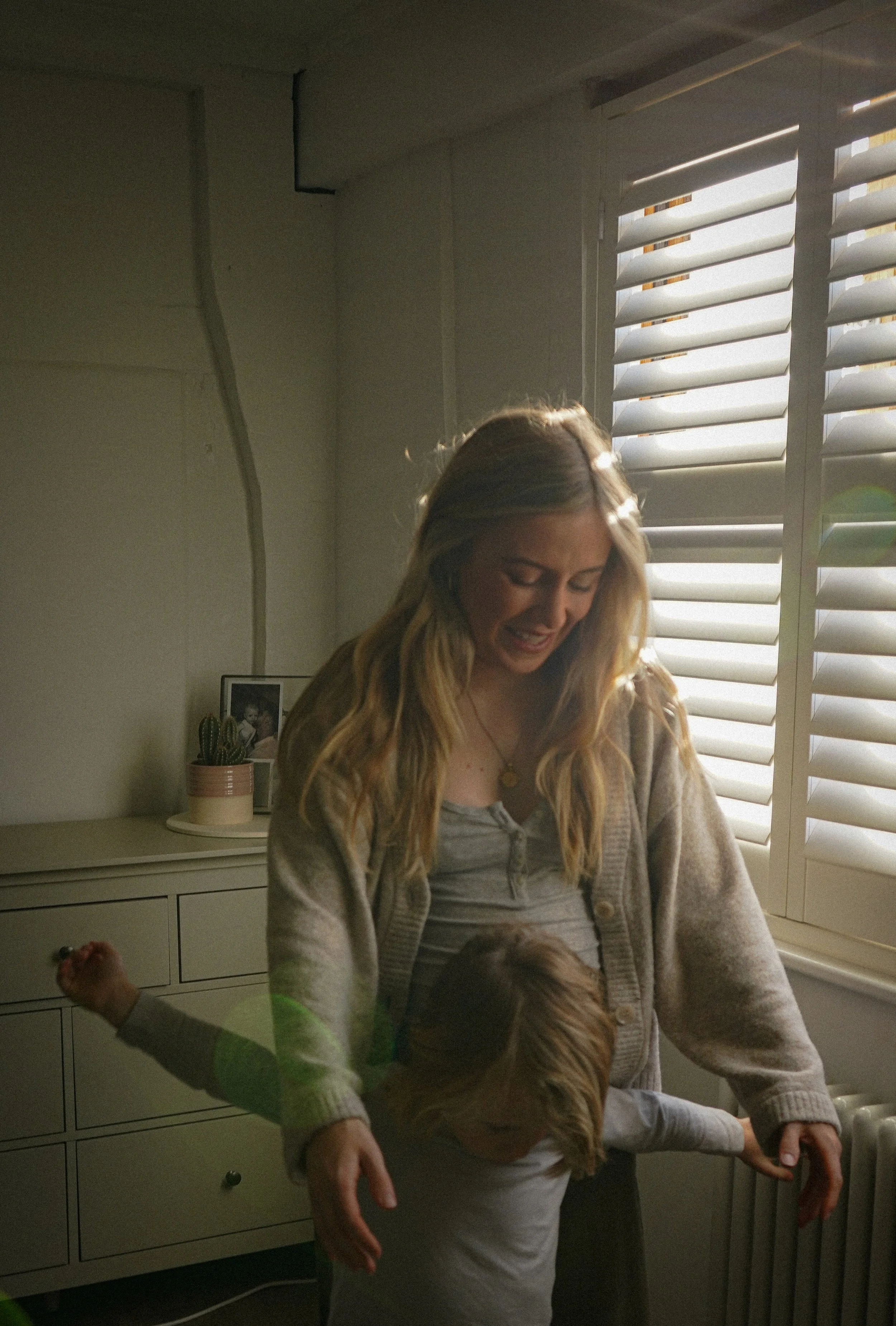 A woman with long blonde hair smiling and looking down, standing near a window with white blinds, wearing a beige cardigan and a gray top, with children playing in front of her.