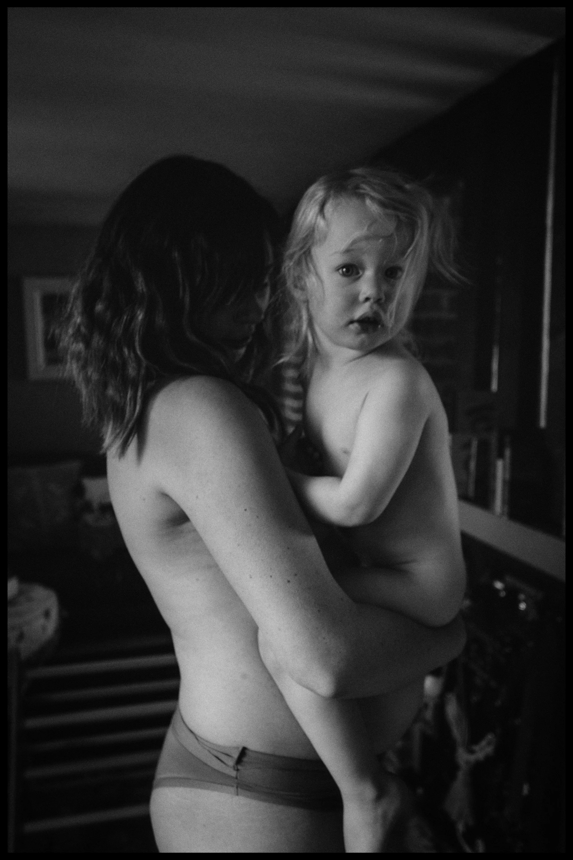 A grayscale photo of a woman holding a young girl, both partially undressed, with the woman facing away and the girl looking directly at the camera. The girl has curly hair and a serious expression, while the woman's face is not visible.