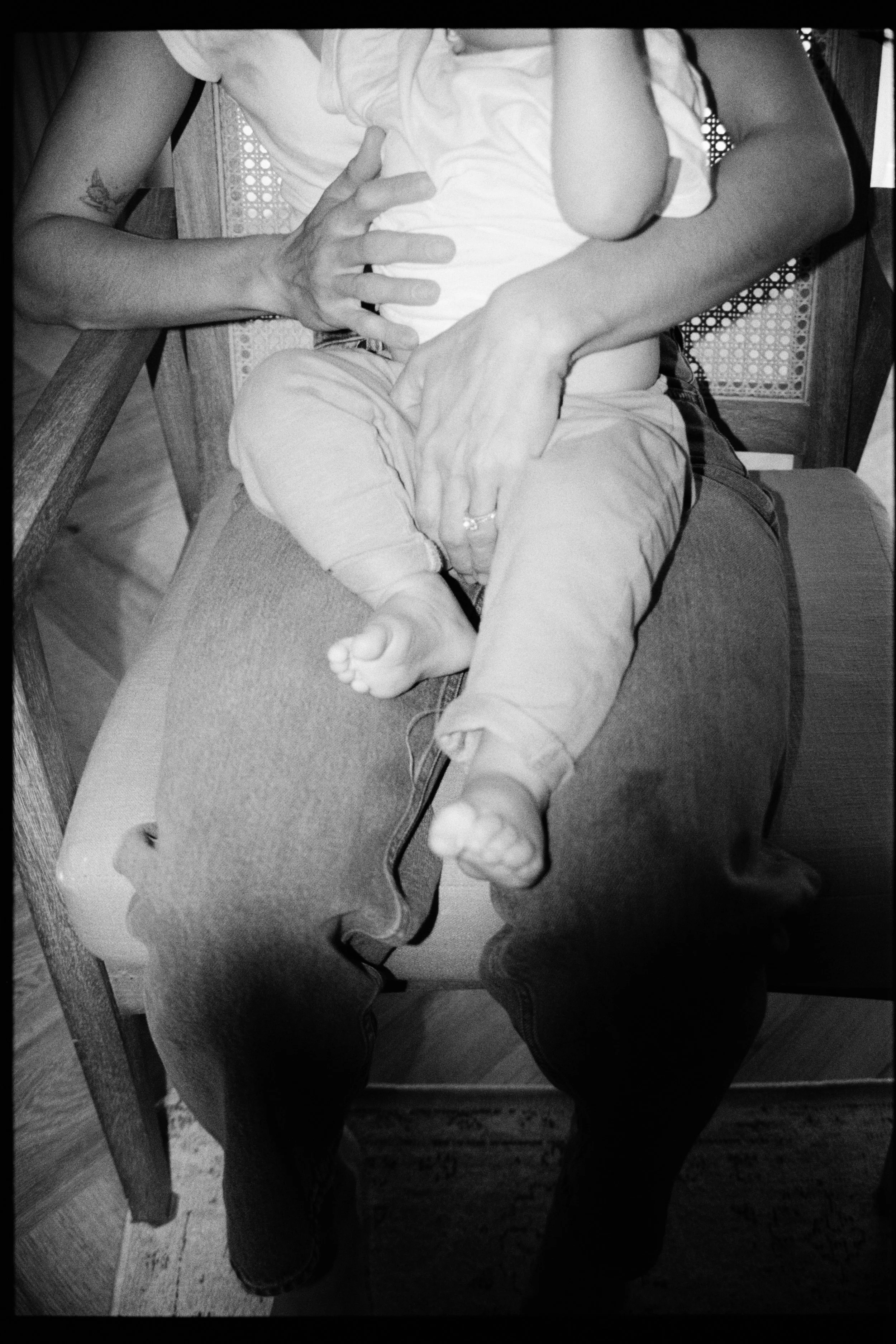 A person sitting on a chair, wearing a white shirt and light-colored pants, with their legs slightly apart. Their right arm is resting on their lap, and their left arm is bent at the elbow, with their hand near their face. A ring is visible on the pe