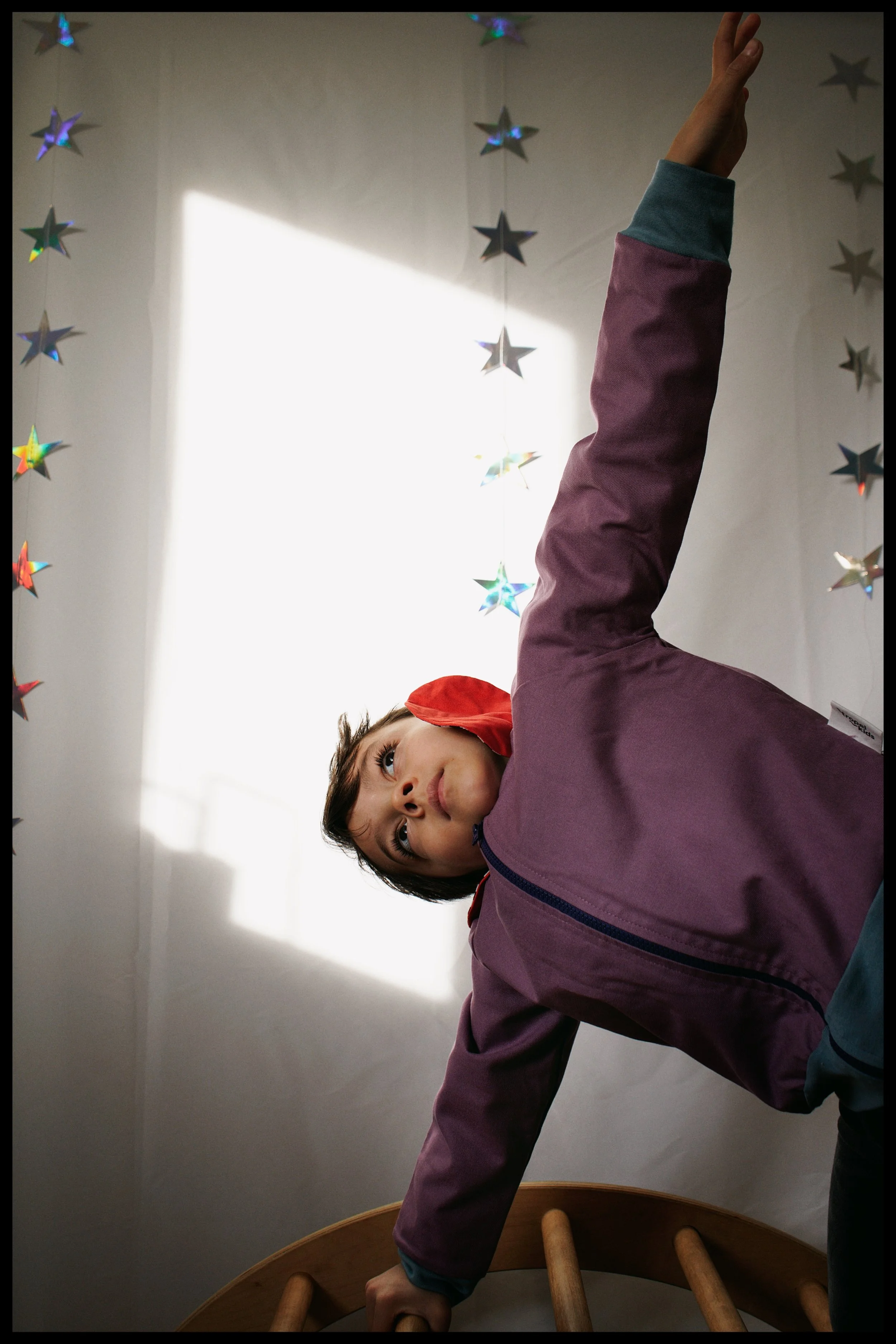 A young boy in a purple jacket and teal shirt, with a red bandana, is seen doing a handstand or balancing on a wooden chair, with his head and arm on the floor. The background includes star-shaped decorations hanging on a wall, and bright light comin