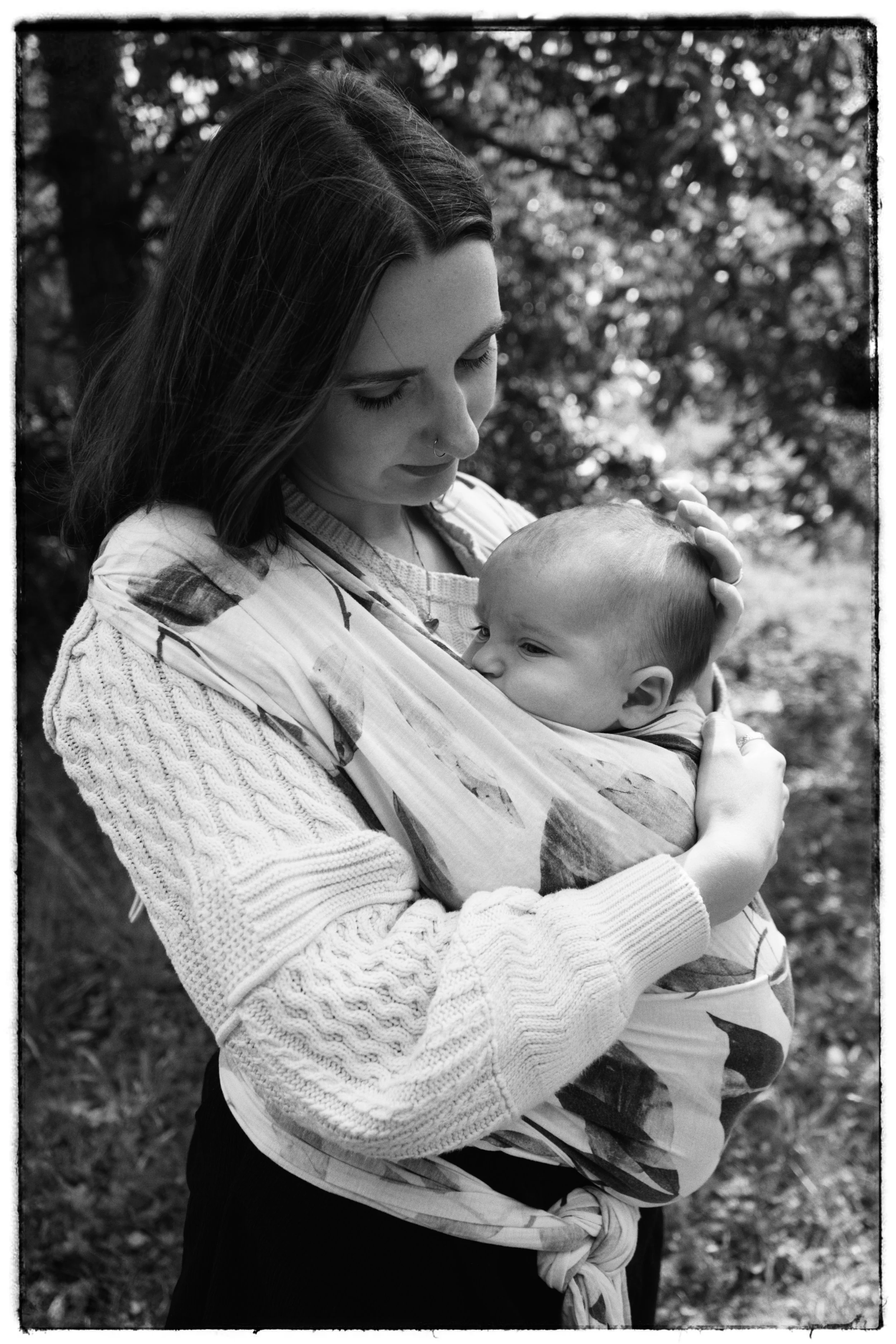 A woman with long hair holding a baby in a wrap carrier outdoors with trees in the background.