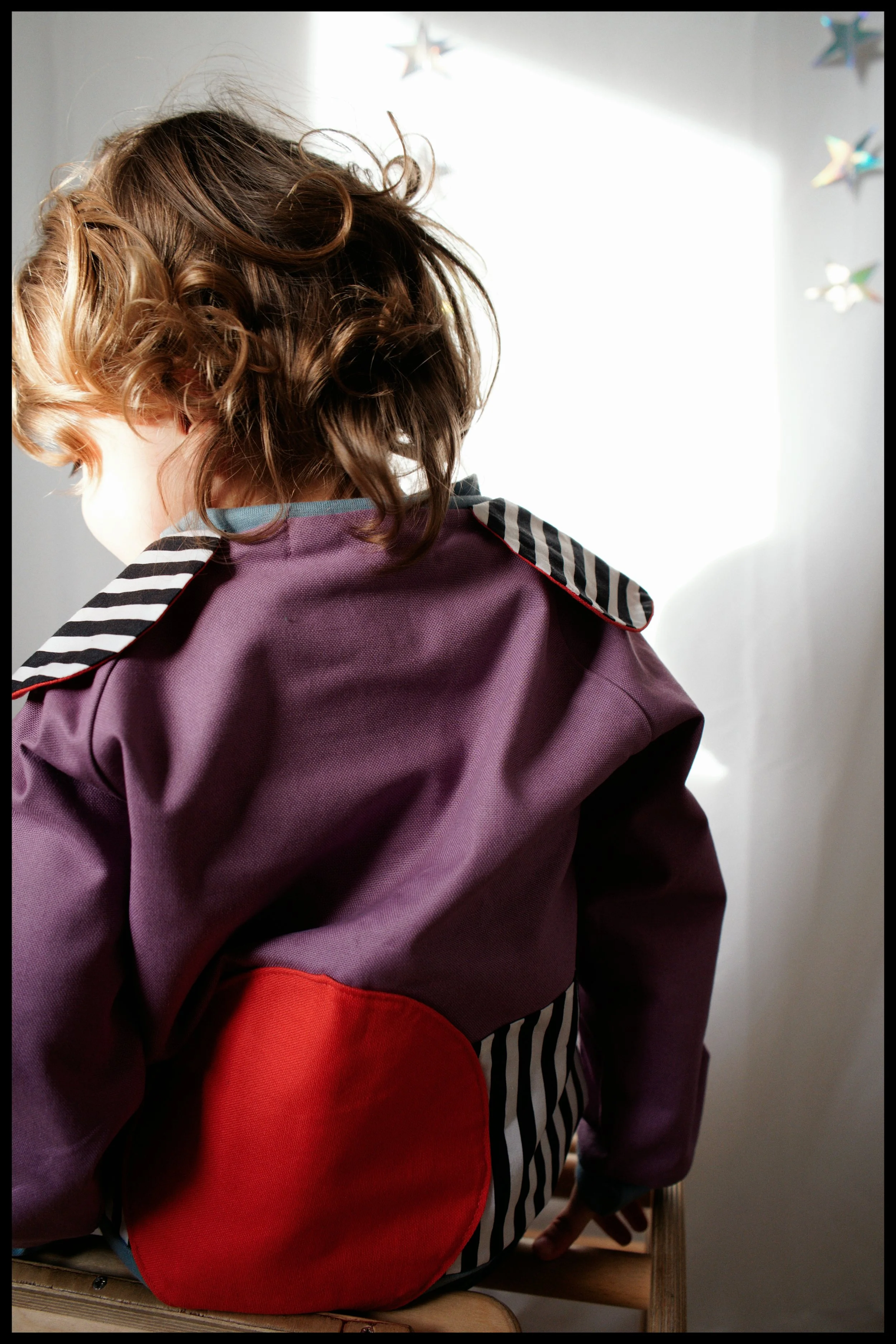 A child with curly brown and blonde hair wearing a purple jacket and a red patch on the back, sitting on a wooden chair, facing away from the camera, near a brightly lit window with colorful paper star and butterfly decorations on the wall.