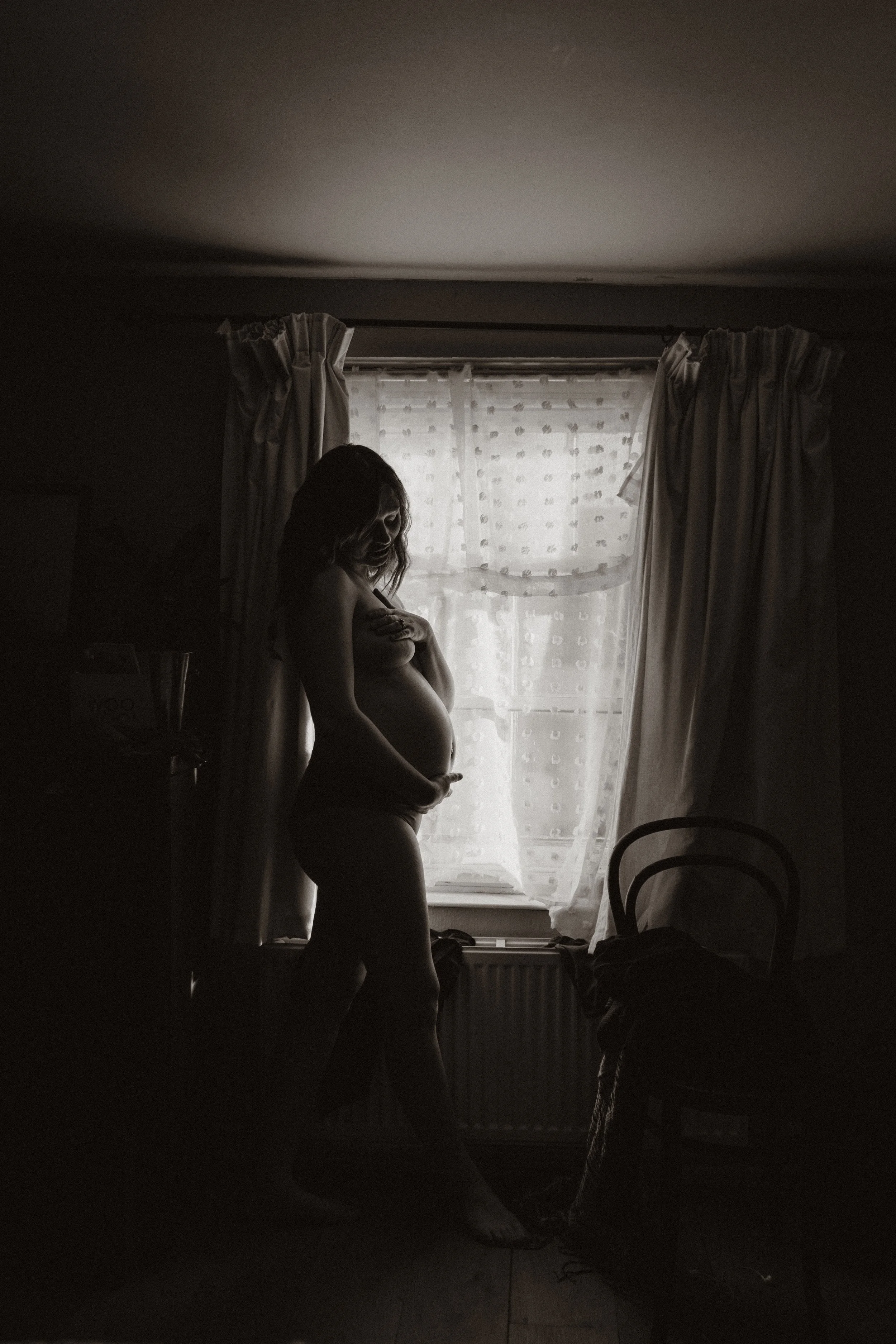 Silhouette of a pregnant woman standing by a window with light curtains, in a dimly lit room.