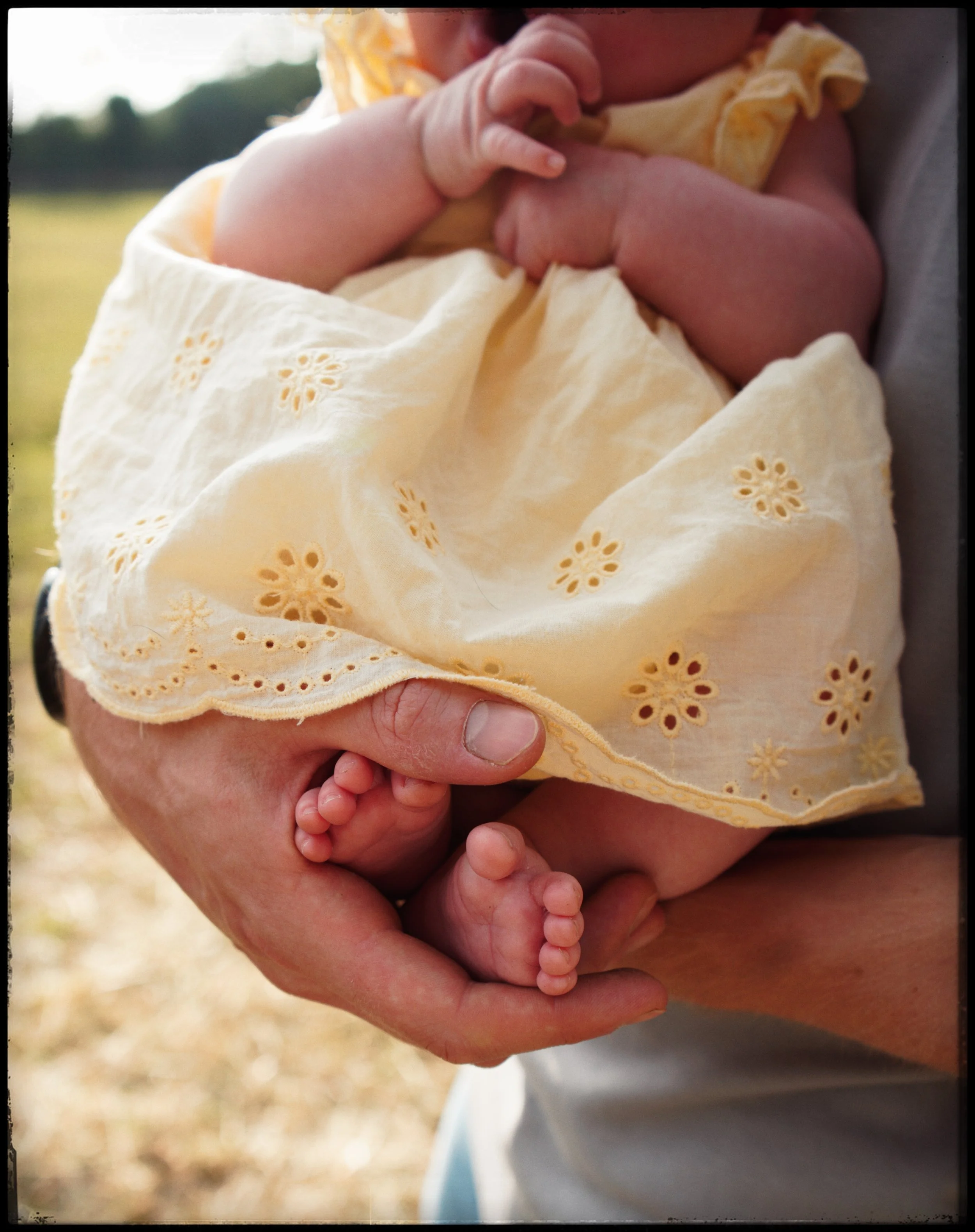 A person holding a baby gently, supporting the baby with one hand around the torso and the other hand holding the baby’s tiny feet. The baby is dressed in a light yellow dress with eyelet embroidery, and appears to be breastfeeding or resting close t