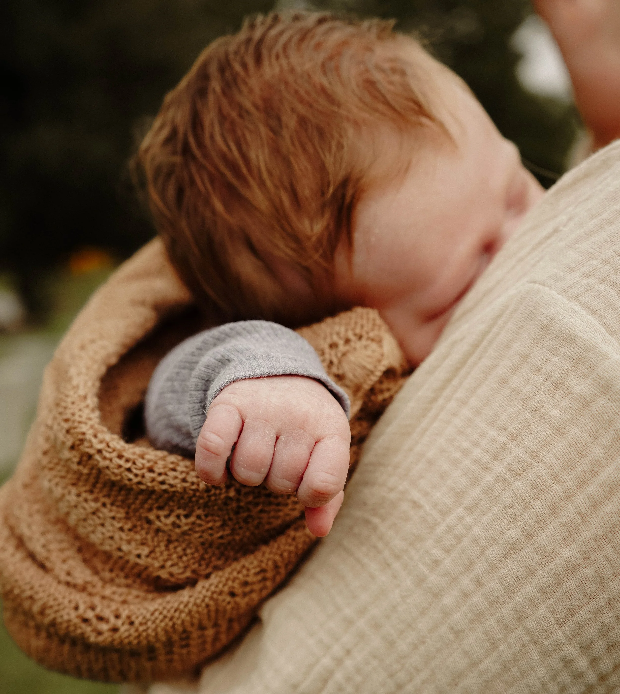 A close-up of a baby with reddish hair, lying on an adult's chest, wearing a gray sleeve.