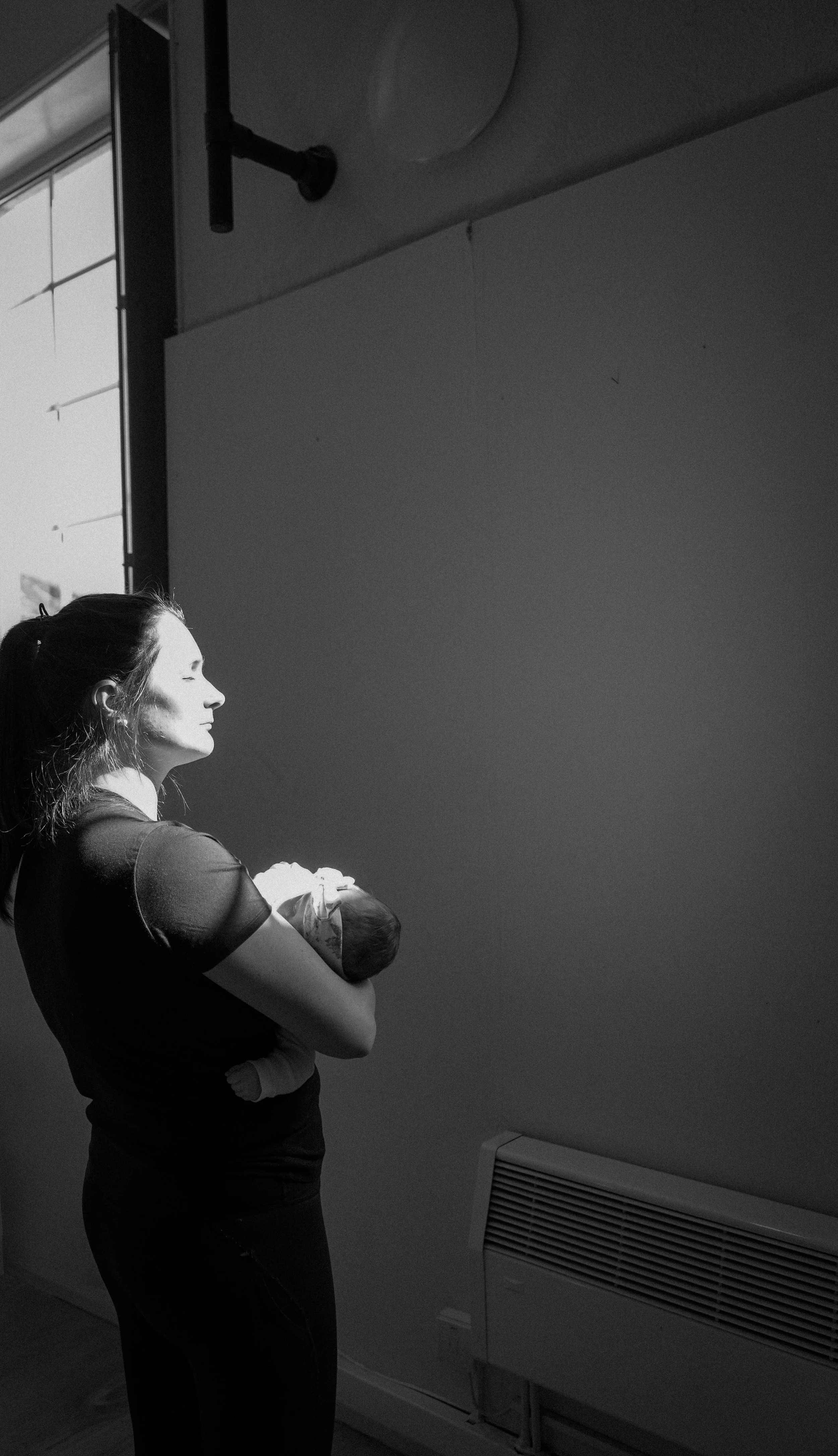 A woman with dark hair in a ponytail holding a baby in a hospital room, with a bright light illuminating her face from the window on the left, and an air vent on the lower right.