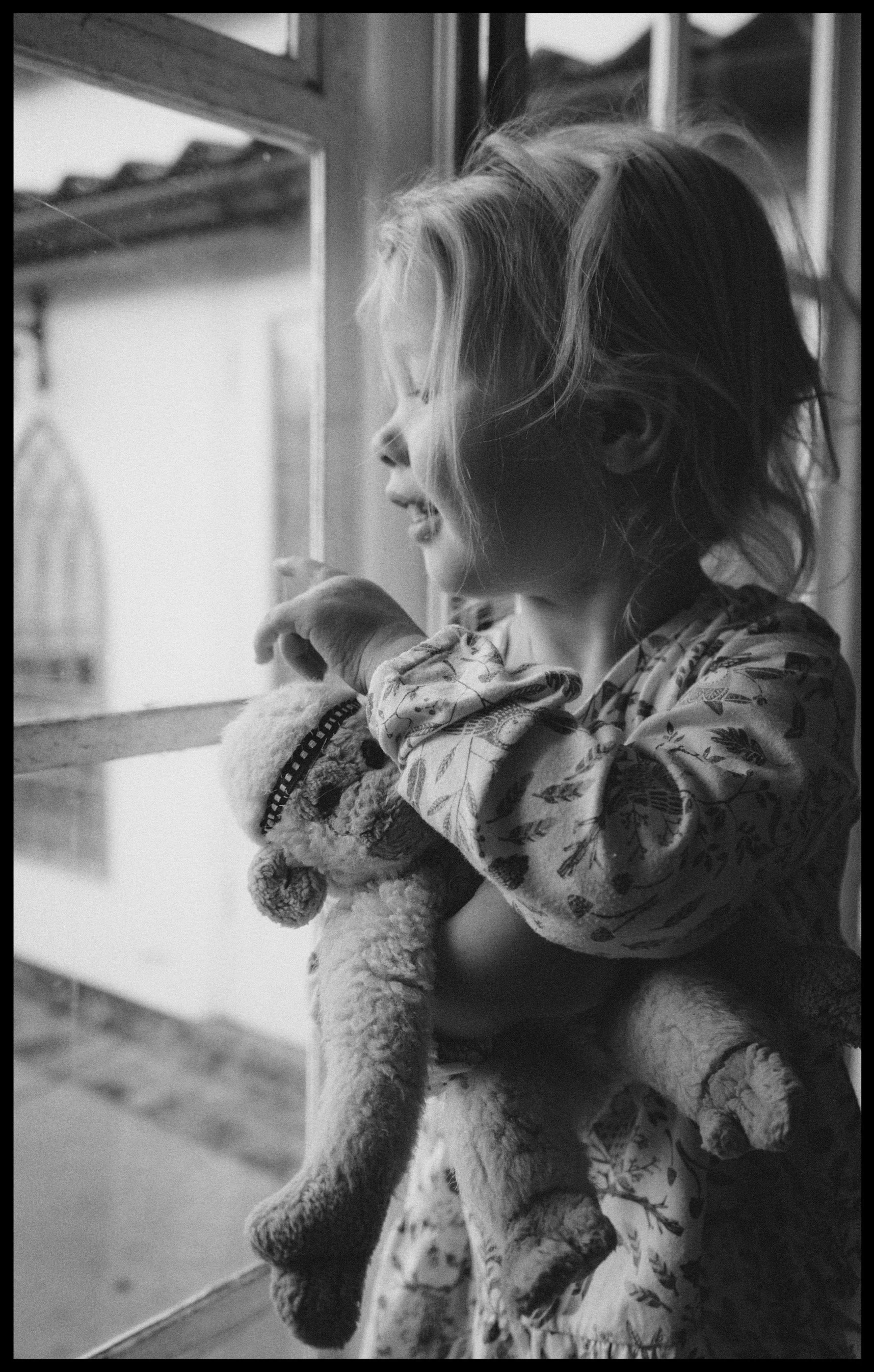 A young child with light hair looking out of a window, holding a plush toy