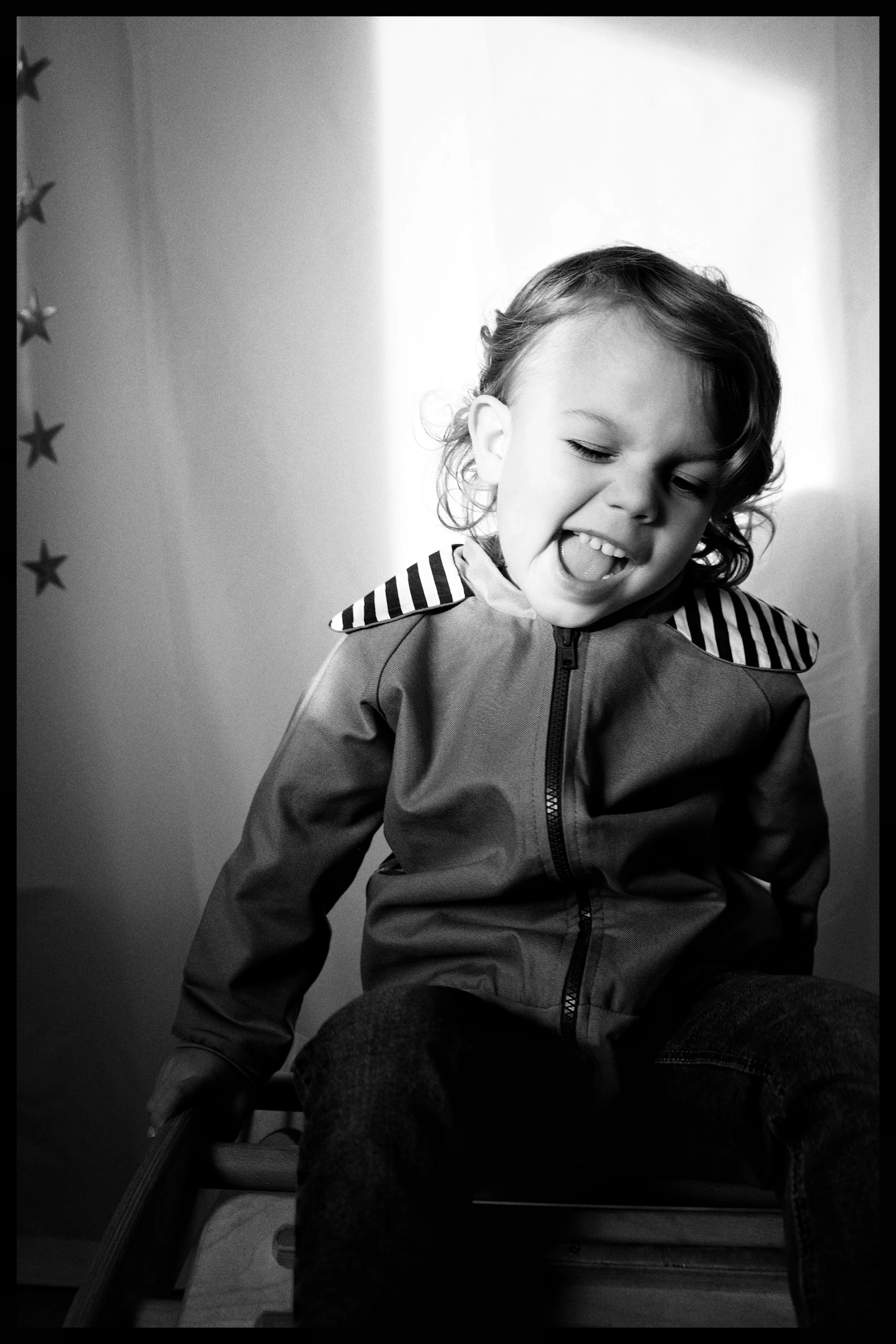 A young girl with curly hair, wearing a jacket with striped shoulder accents, is sitting on a chair with her eyes closed and mouth open, appearing to be singing or speaking. The background shows a wall with star-shaped decorations.