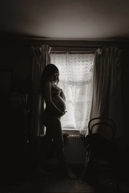 Silhouette of a pregnant woman standing by a window with curtains, holding her belly in a dimly lit room.