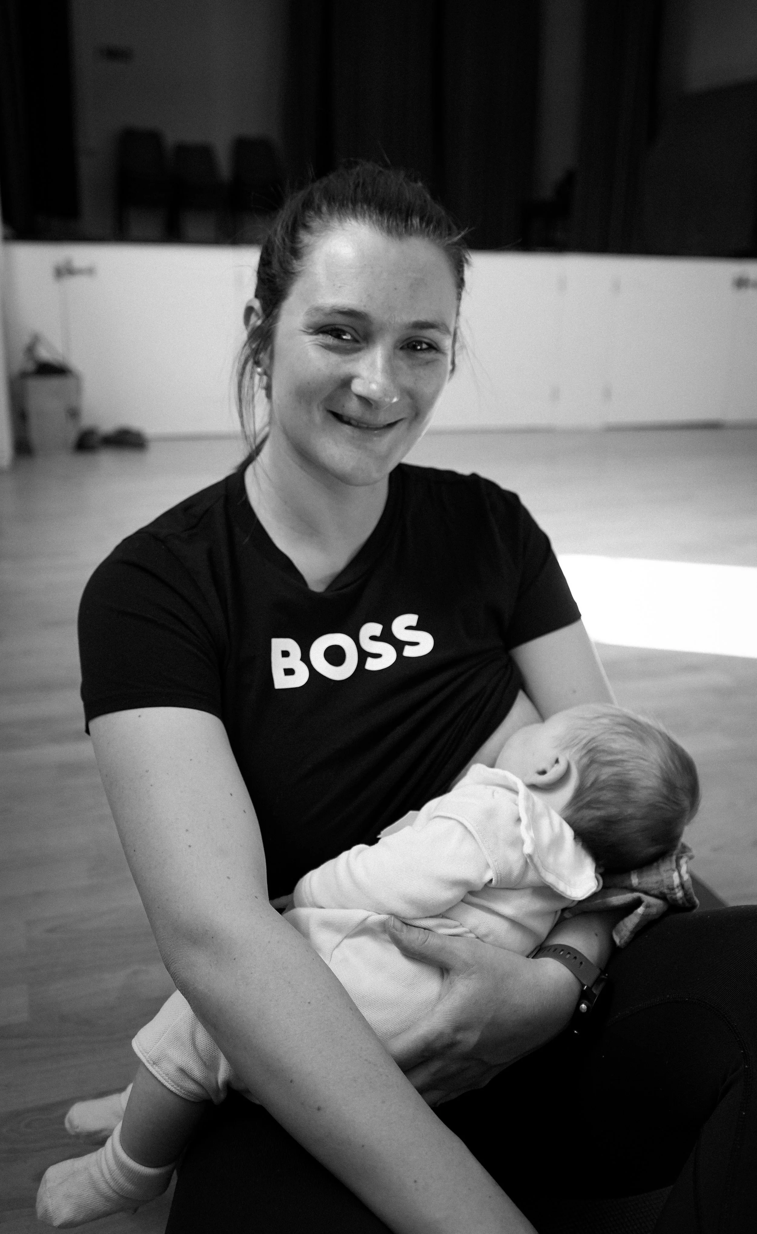 A woman with dark hair in a ponytail smiling and holding a sleeping child in her arms. The woman is wearing a black t-shirt with the word 'BOSS' printed on it. The background shows a room with some chairs and a radiator.