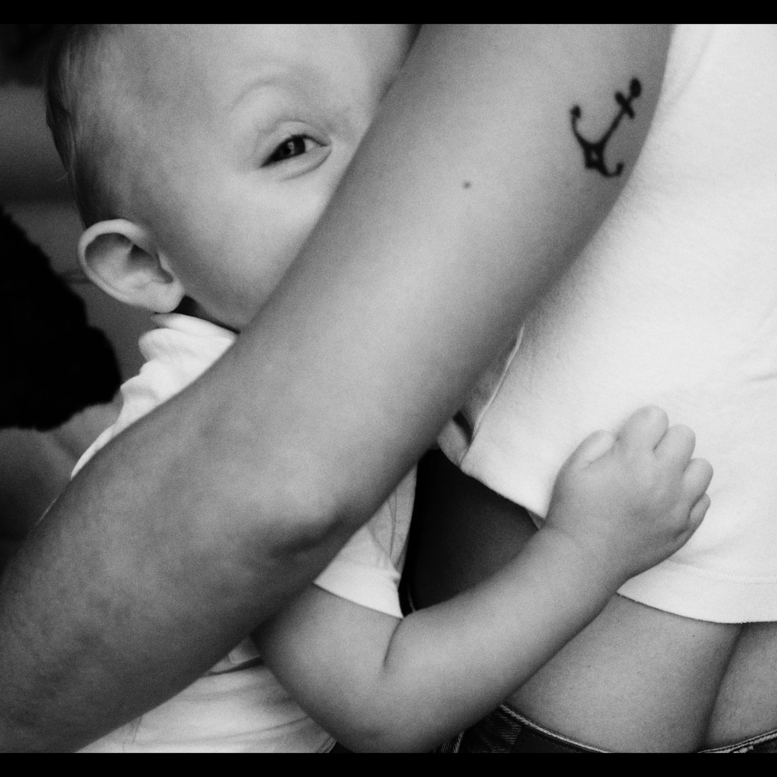 Close-up black and white photo of a woman’s face, showing her eye, ear, and a tattoo of an anchor on her upper arm. She is resting her chin on her hand, with her fingers near her lips.