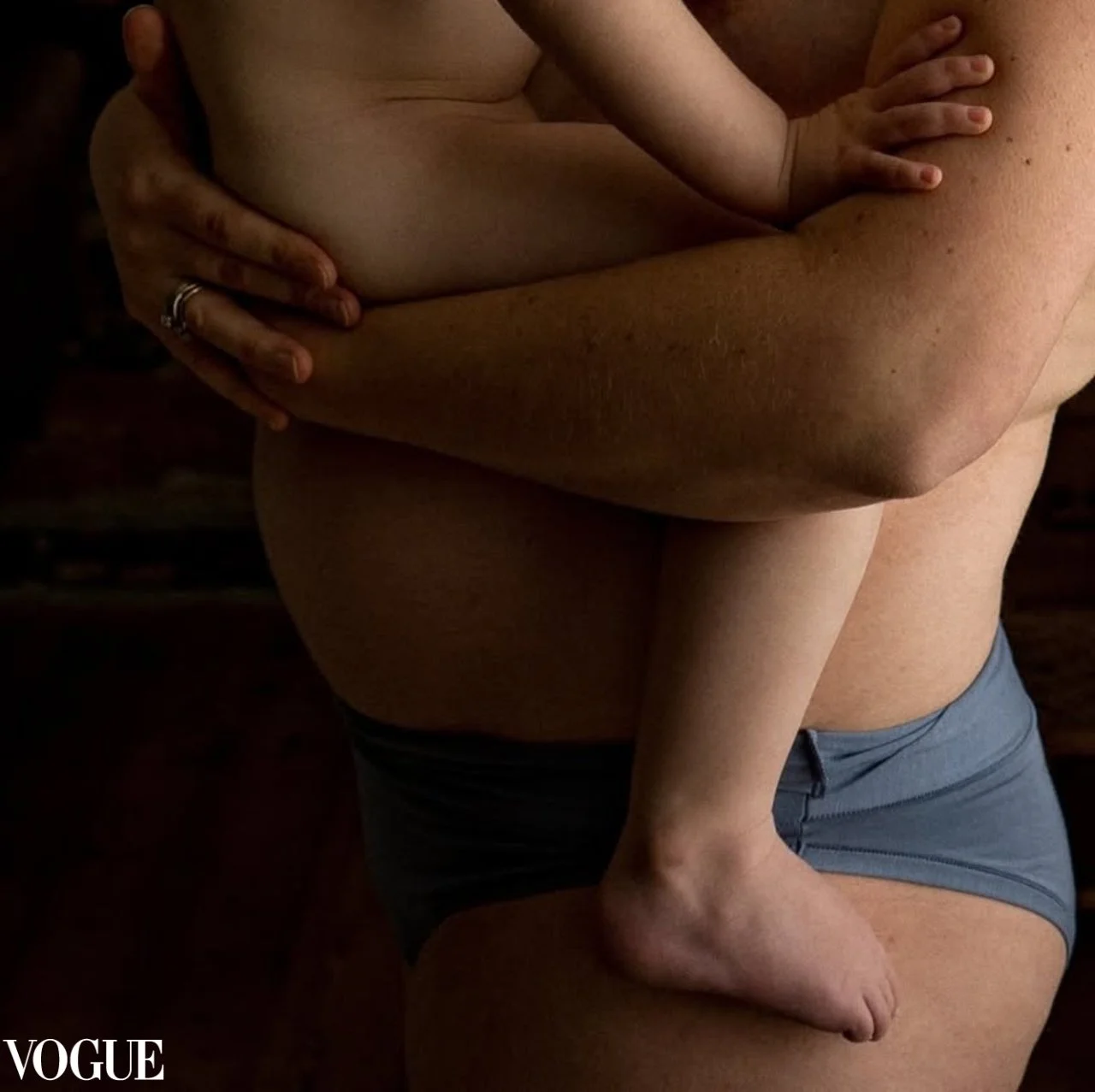 A close-up of a person wearing blue shorts, holding a child with their arms crossed in front of their chest. The child's legs are wrapped around the person's waist, and the child's hand rests on the person's shoulder. The background is dark and out o