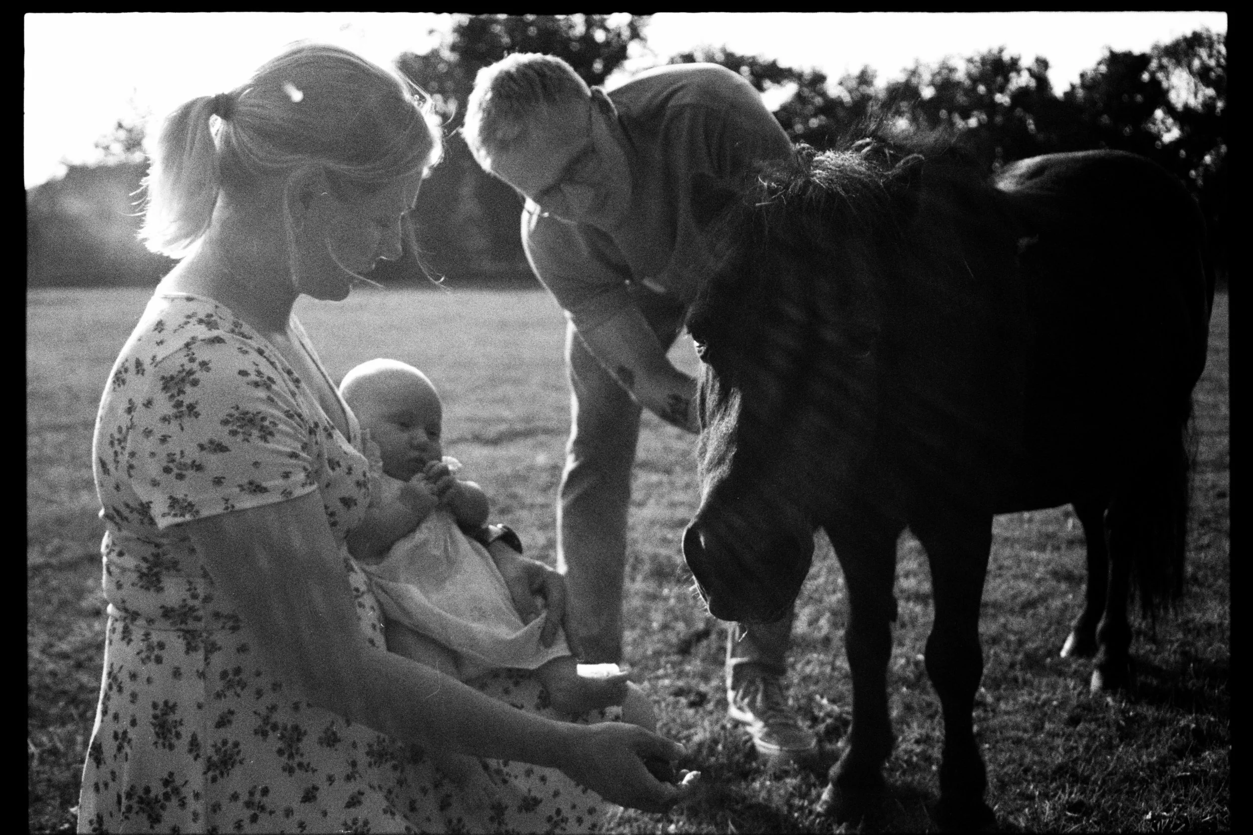 A black and white photograph of a woman holding a small child, with a man and a horse nearby outdoors with trees in the background.