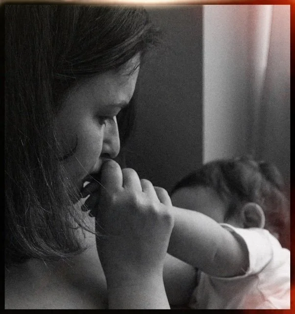 A woman with shoulder-length hair holding a small child's arm or hand in her hands, appearing to be thinking or reflecting with a serious expression, in a dimly lit room.