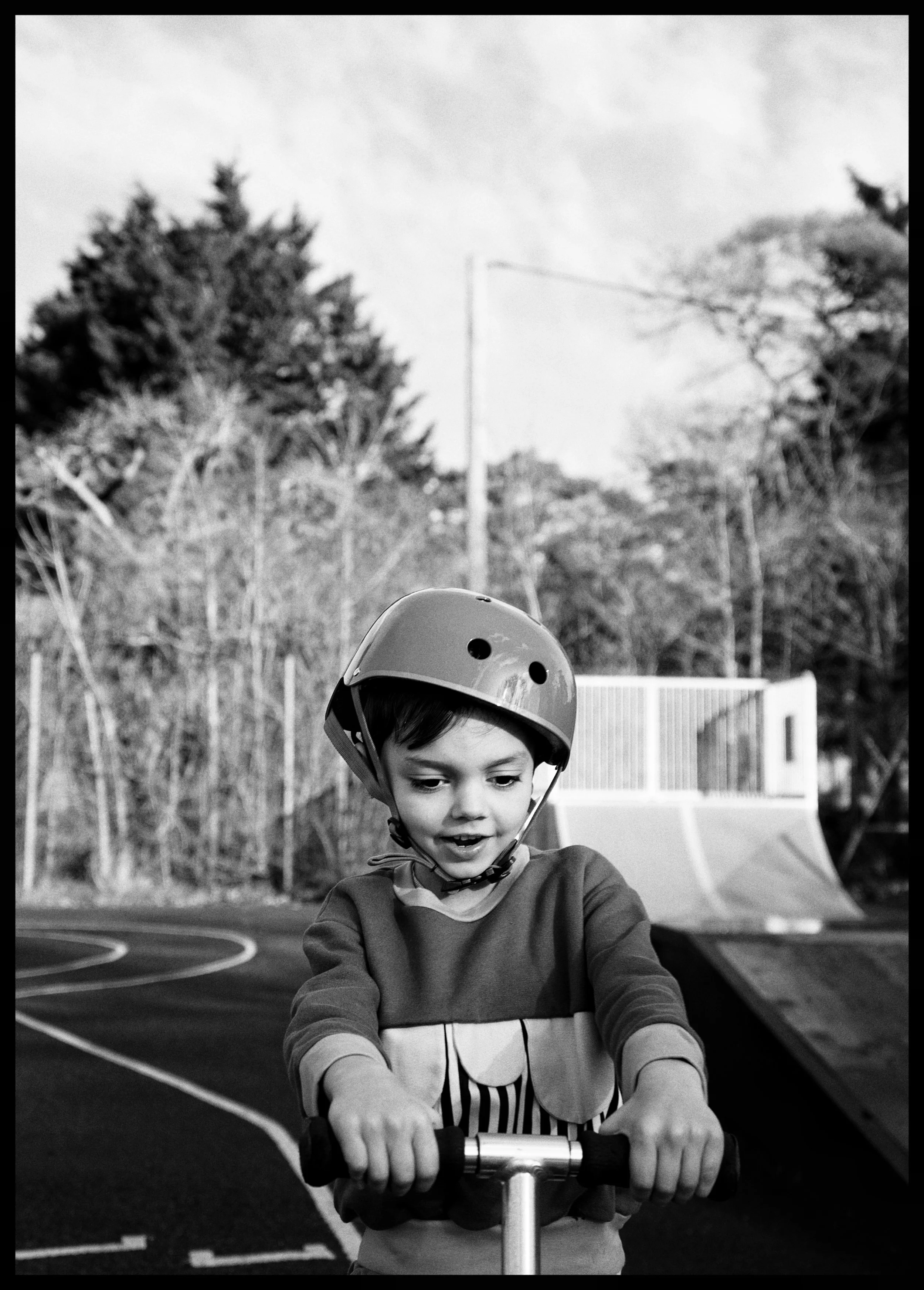 A young boy wearing a helmet riding a scooter or bike on a sports court, with trees and a building in the background.