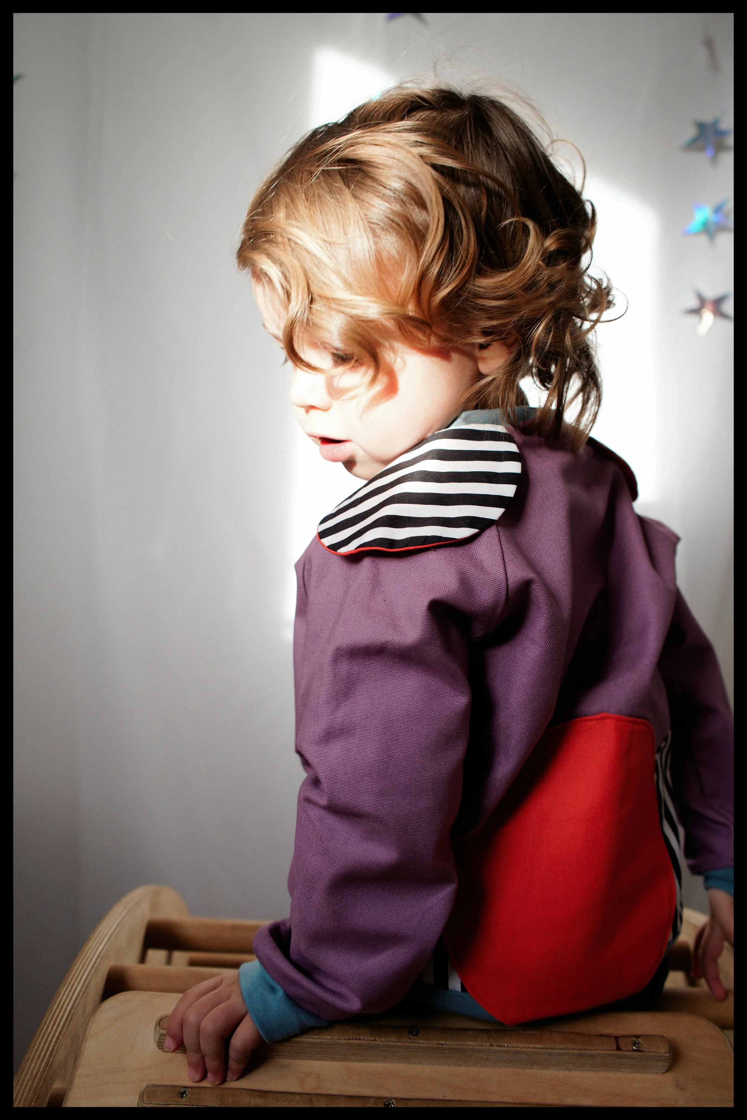 A young child with curly hair, wearing a striped shirt, a purple jacket, and red pants, sitting on a wooden bench or playtrain