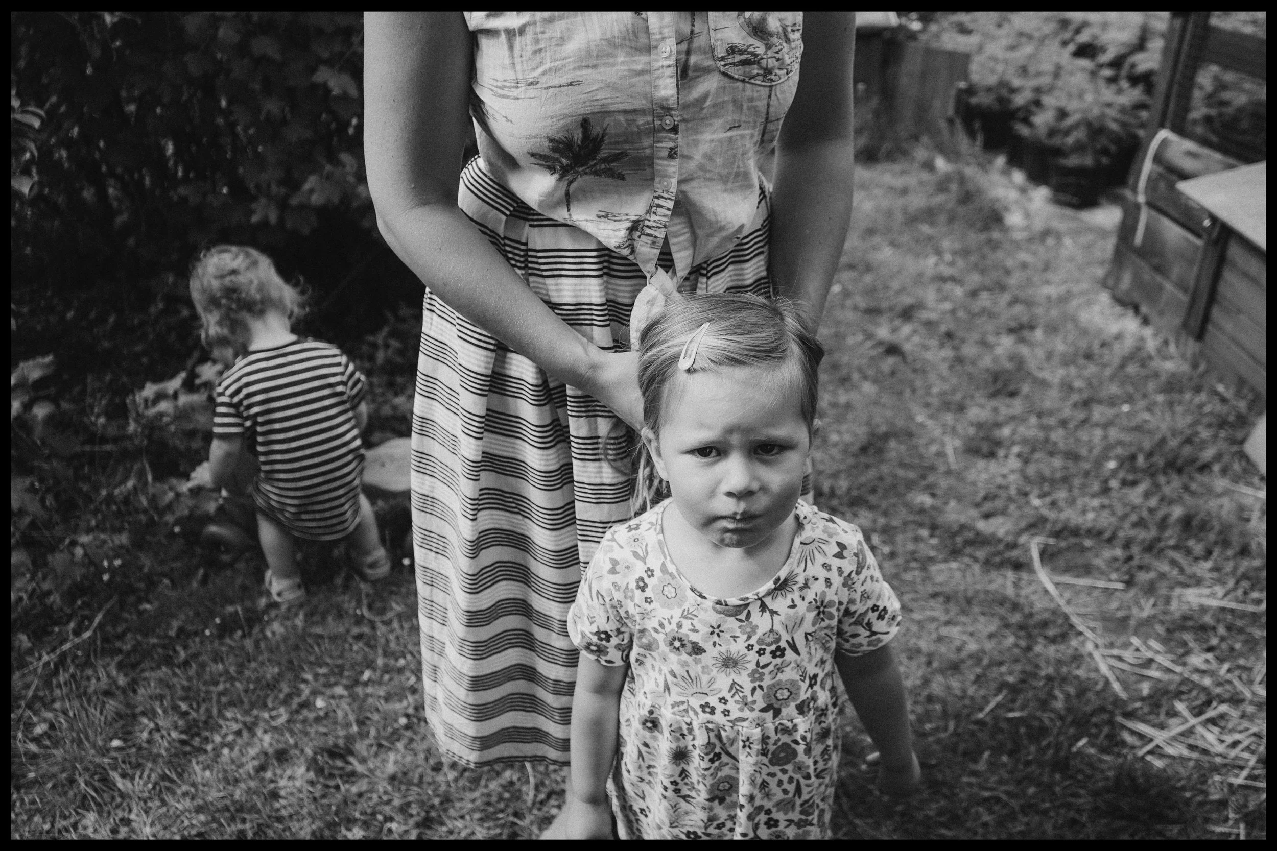 A young girl with a serious expression stands outdoors in a floral dress, with her hair held back by a hairpin, while an adult in a summer dress with a palm tree pattern holds her shoulders. In the background, a child in a striped shirt is sitting on