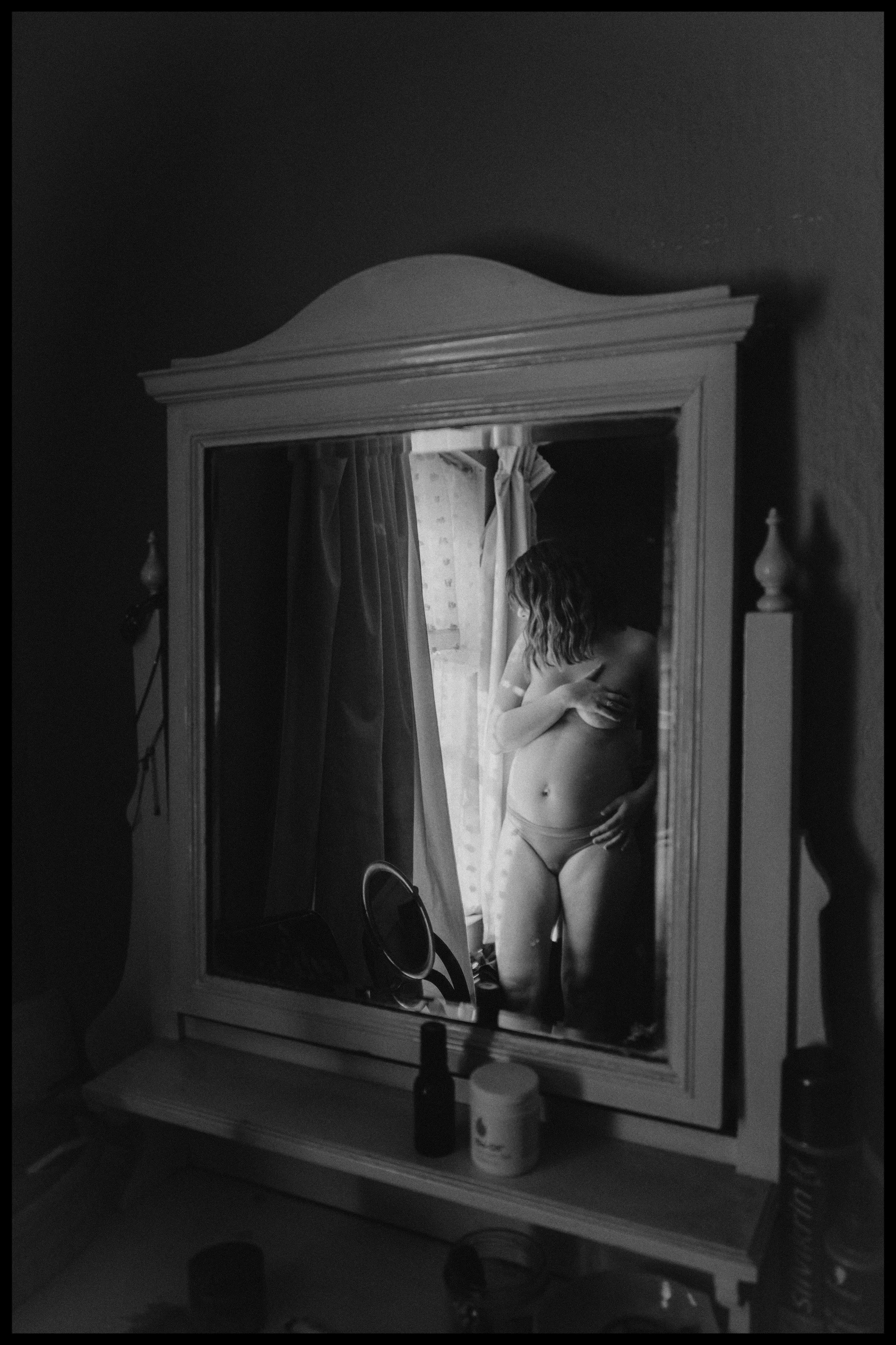 A woman stands in front of a mirror topless, wearing underwear, with her arms crossed over her chest. The mirror shows her reflection in a room with curtains and a plain wall. The photo is in black and white.