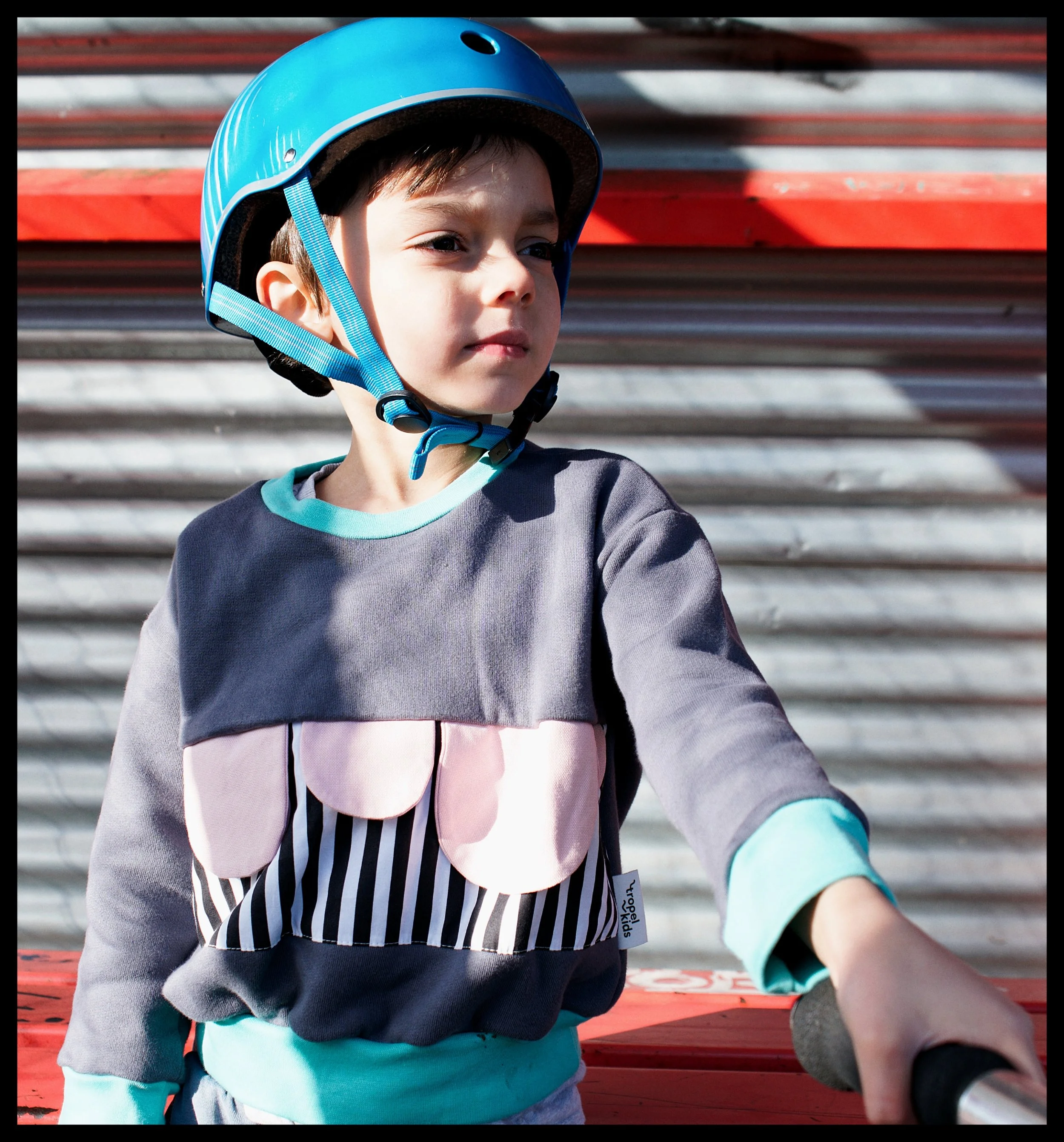 Close-up of a young boy wearing a blue helmet and colorful jacket, sitting on a red park bench.