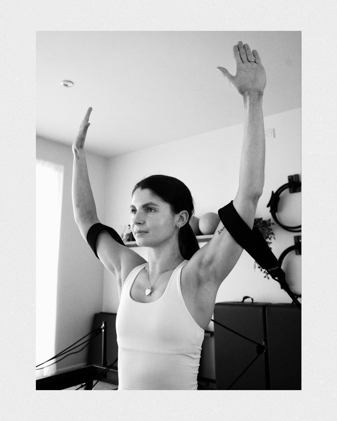 A woman with dark hair tied back, wearing a tank top, standing indoors with her arms raised overhead and black resistance bands around her upper arms.