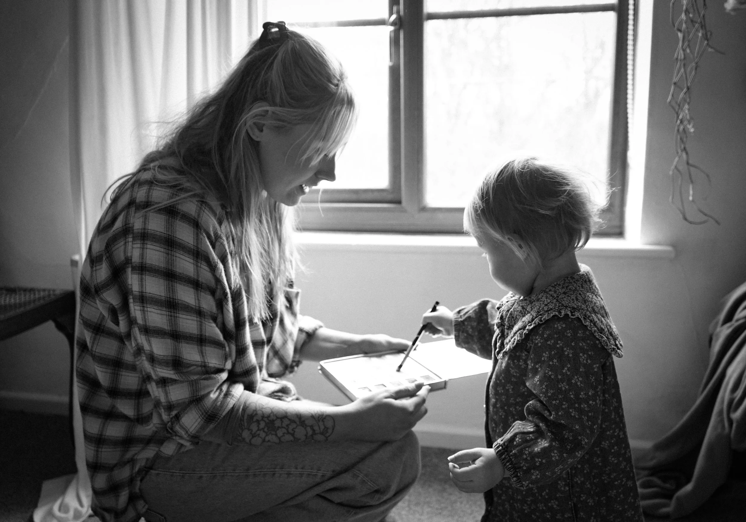 A woman with long hair and a checkered shirt is sitting and smiling while a young girl with short hair in a patterned dress stands in front of her. The woman is holding a tablet and showing it to the girl, who is holding a stylus. They are in a brigh