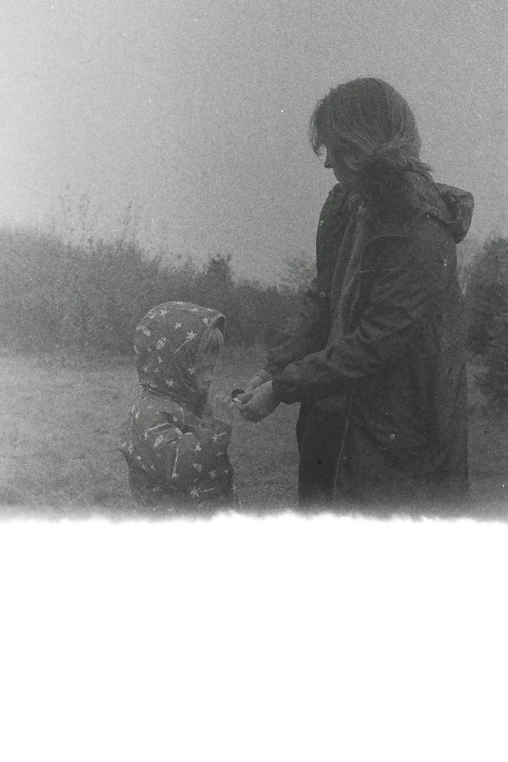 A woman in a jacket and a child in a star-patterned hoodie standing outdoors, with the woman handing something to the child during dusk or foggy weather.
