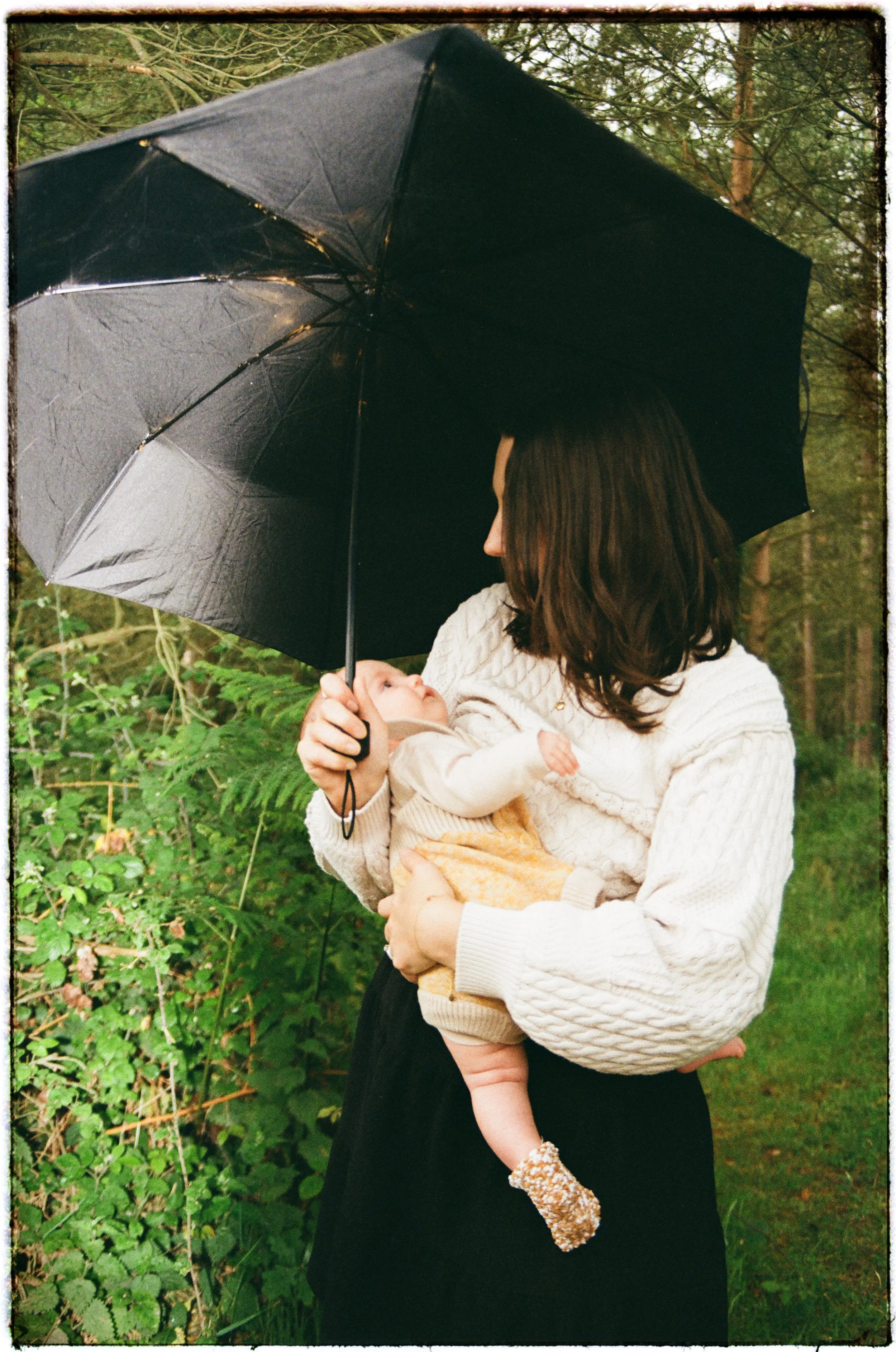 A woman holding a baby outdoors under a black umbrella, surrounded by greenery and trees.