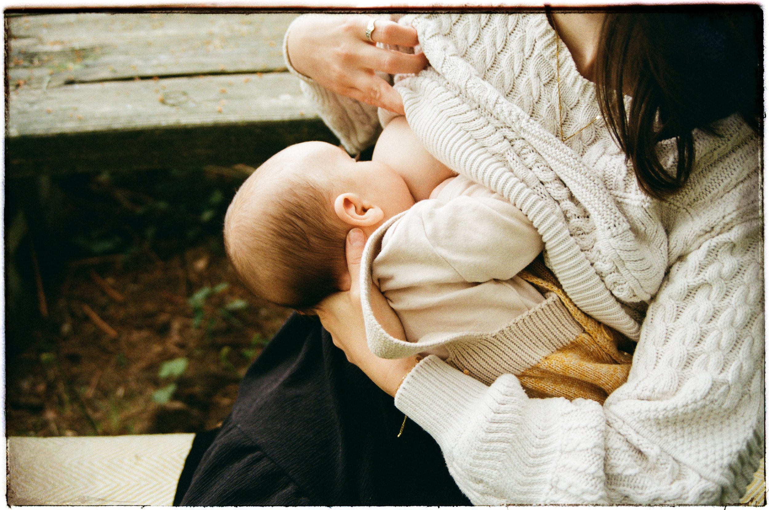 A person holding a sleeping baby in their arms, sitting outdoors on a wooden bench, with earthy ground and leaves visible in the background.