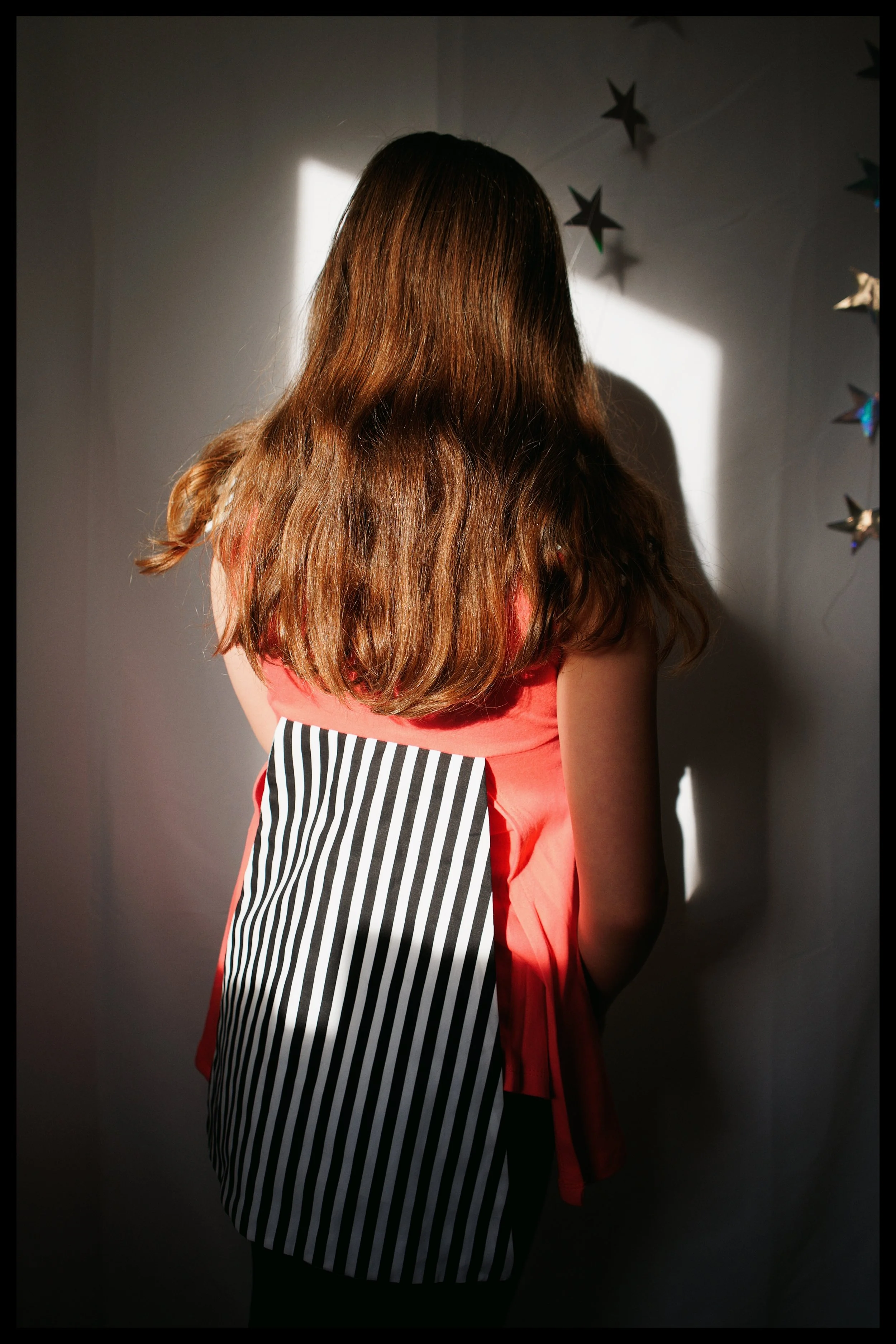 Back view of a woman with long wavy brown hair, wearing a red dress with a black-and-white striped apron, standing in front of a neutral wall decorated with holographic star-shaped ornaments. A beam of light creates a shadow on the wall.