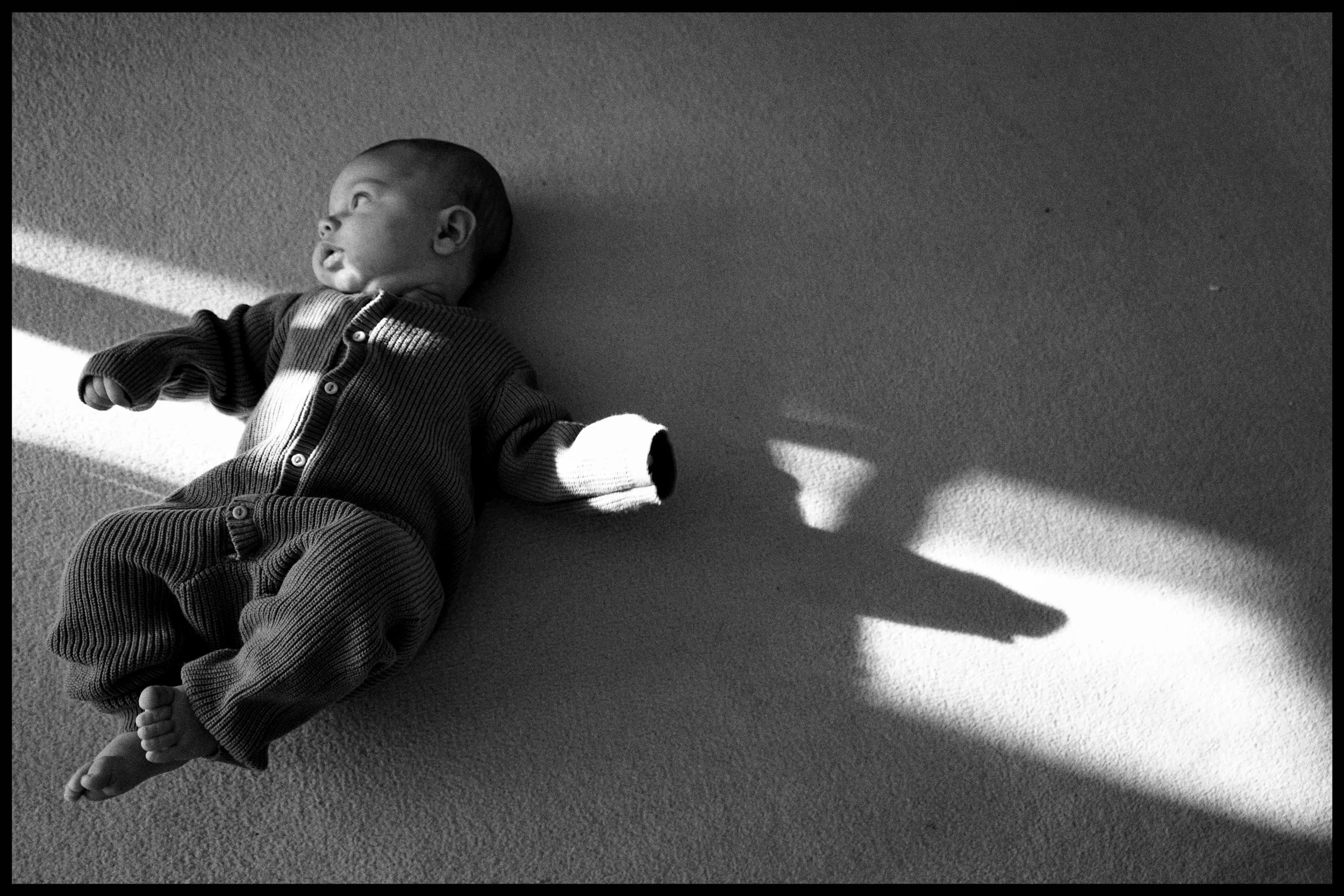 A young child lying on a carpeted floor, looking to the side, with patterned clothing, sunlight casting shadows on the floor.