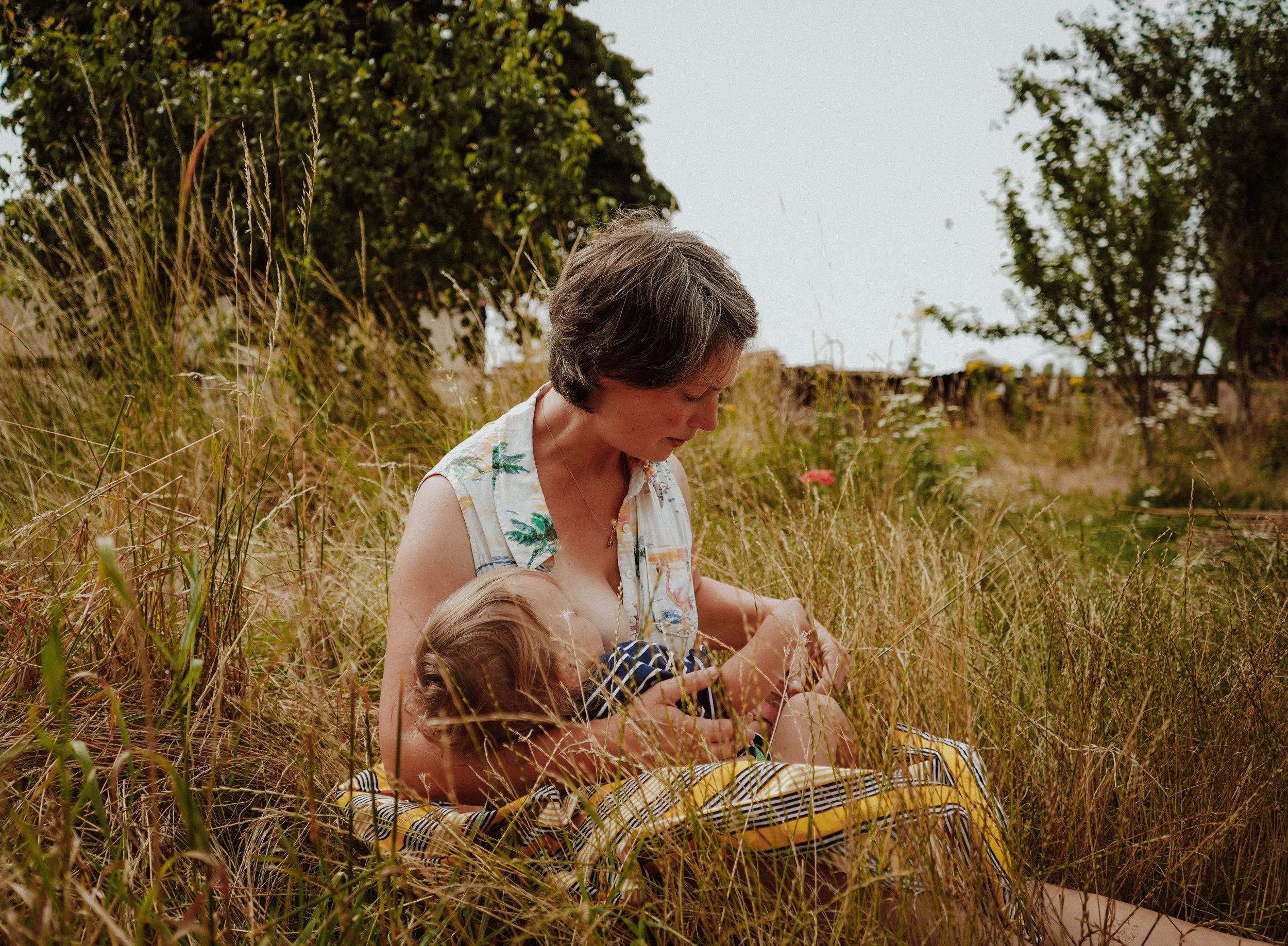 A woman and a young girl with blonde hair sitting in a field of tall grass, surrounded by trees, during daytime.