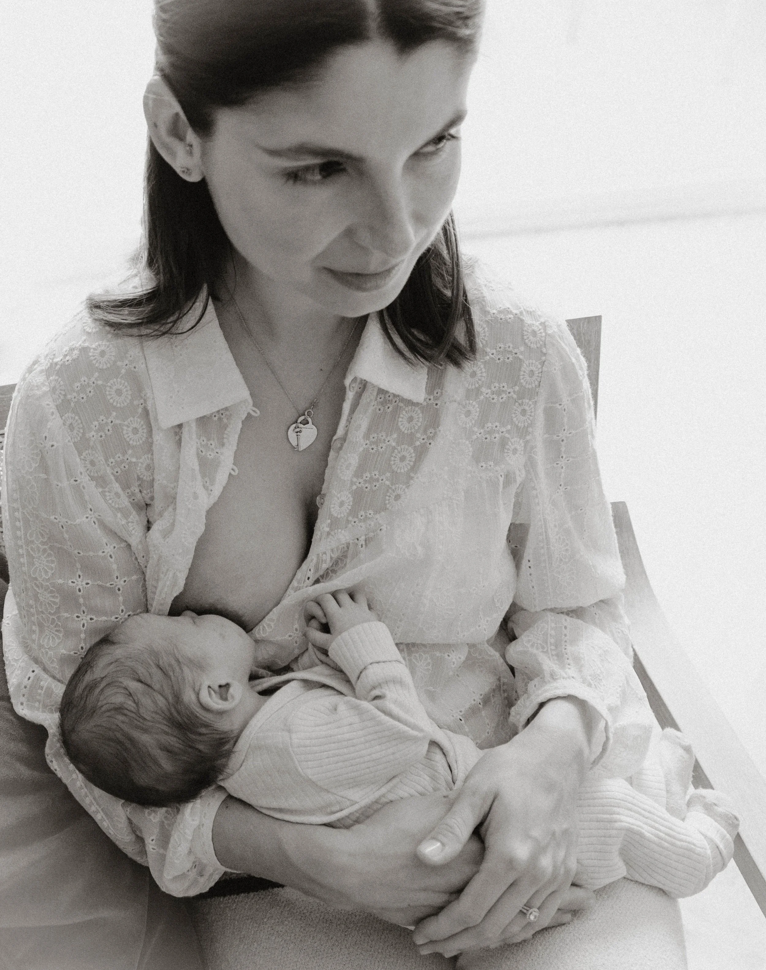 A woman with dark hair and earrings gently cradles a sleeping newborn baby in her arms. The woman is sitting, wearing a white, embroidered top, with a necklace featuring a heart-shaped pendant. The baby is dressed in light clothing and appears peacef