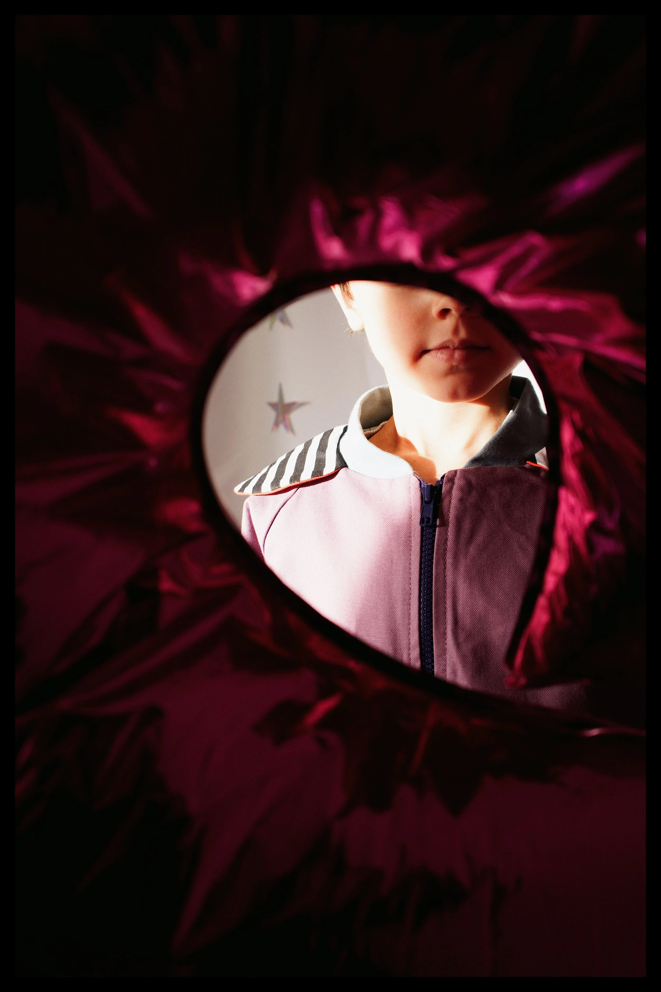 A young girl looking through a heart-shaped opening in a red and pink object, with a star-shaped decoration hanging in the background.