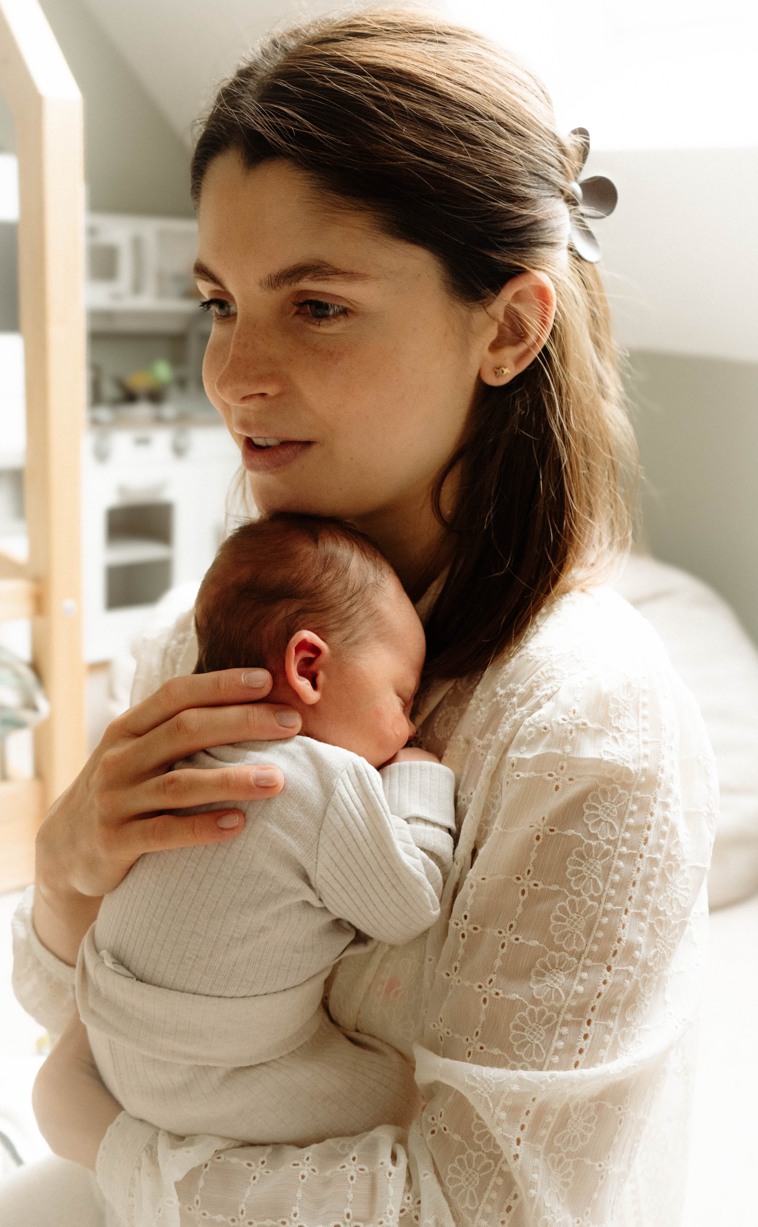 A woman with brown hair holding a sleeping baby close to her chest indoors, with a soft background.