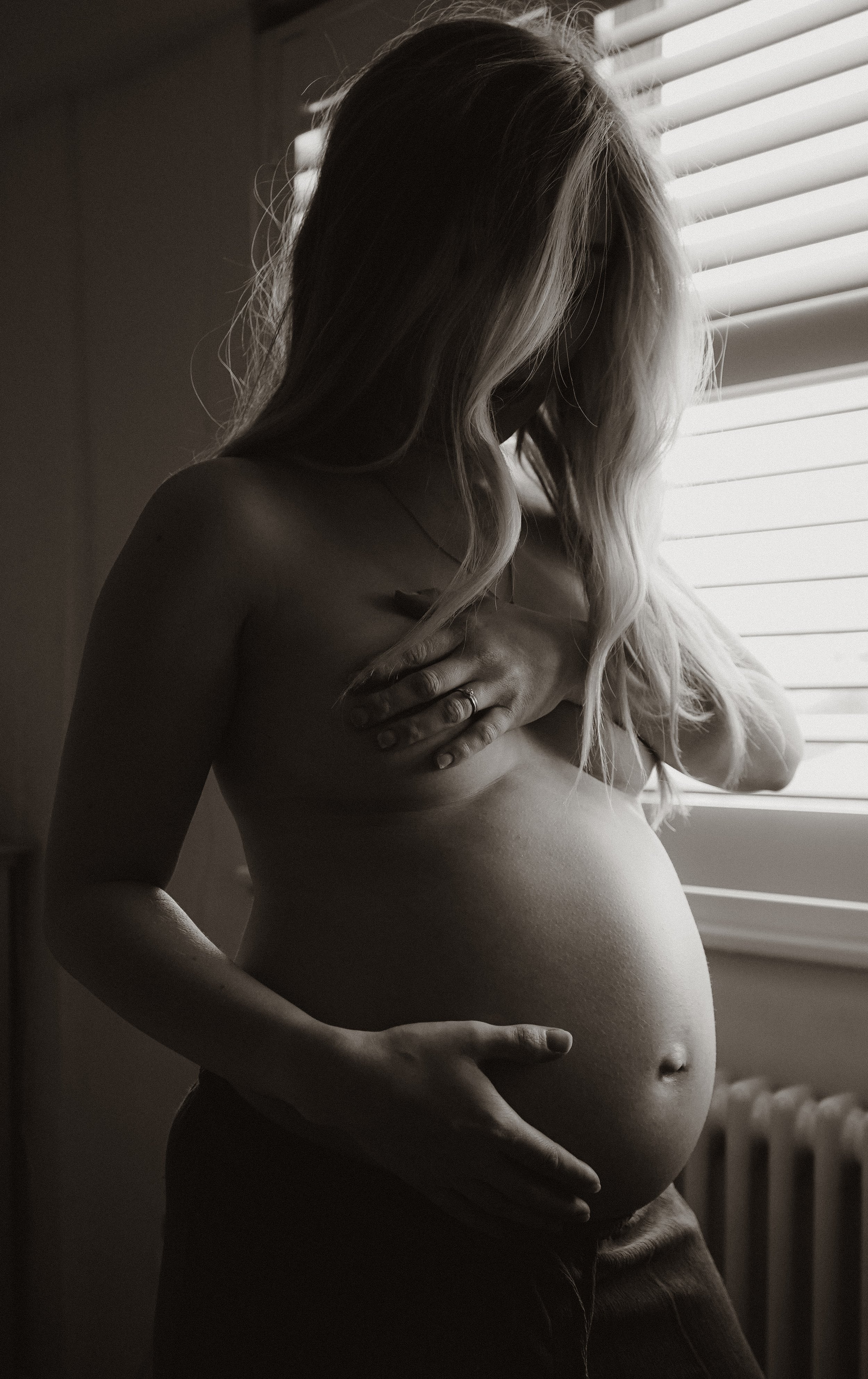 A pregnant woman with long hair standing near a window with closed blinds, touching her belly and chest.